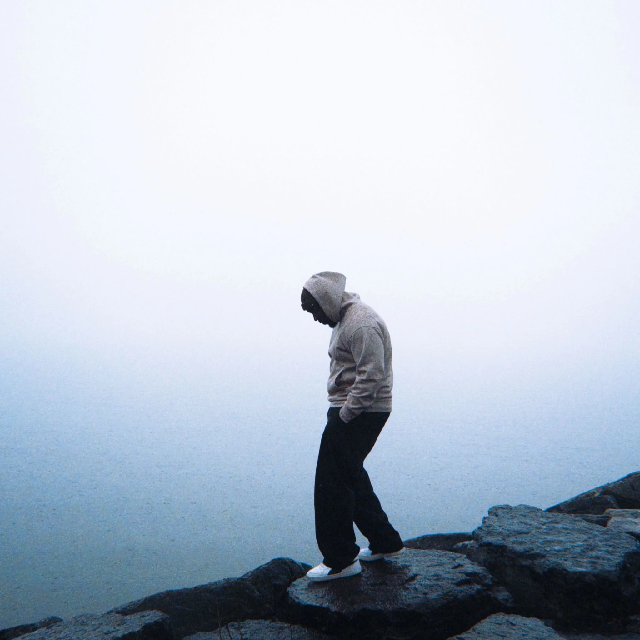 Kojo Dave standing on rocky shoreline near water during overcast weather with a foggy sky.