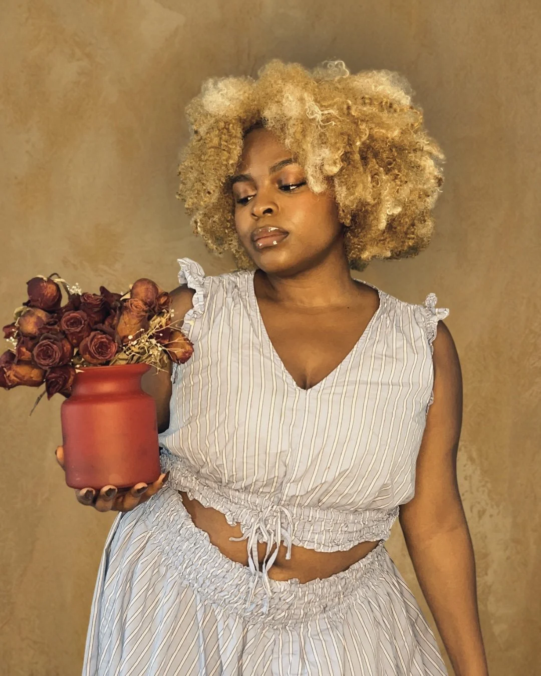 DĀHNI with curly blonde hair holding a red vase with dried roses, looking down.