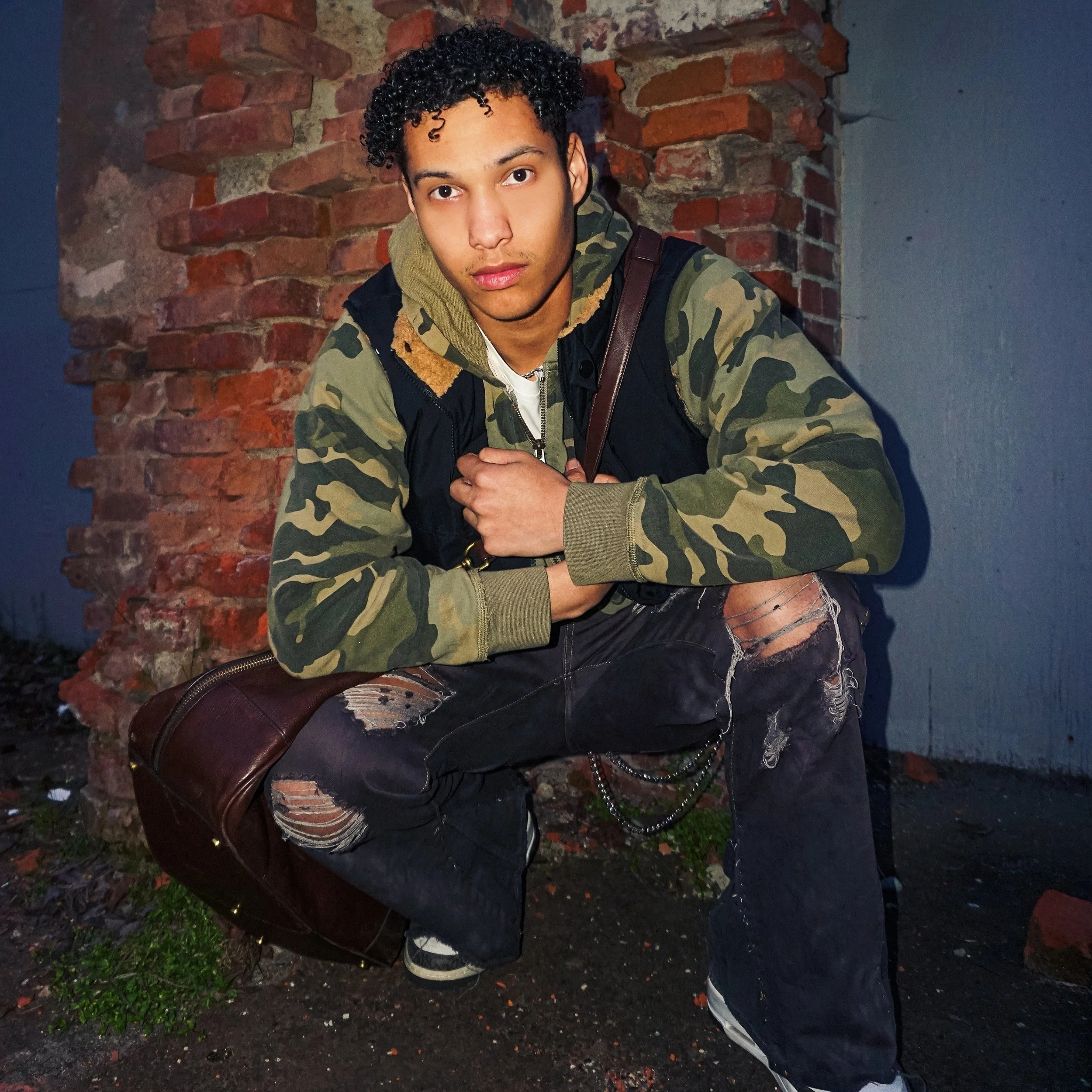 Jalen Jordan sitting on a bench with a brick wall behind him, wearing a camouflage jacket and ripped jeans, with a brown bag on his lap, looking directly at the camera.
