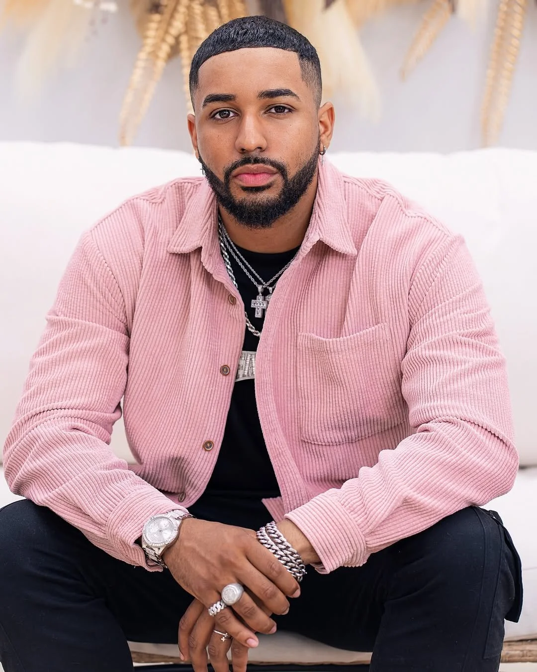 Fame Holiday with a well-groomed beard, wearing a pink corduroy shirt over a black T-shirt, sitting on a light-colored sofa with a blurred background and decorative plants, accessorized with jewelry, including chains, rings, a watch, and bracelets.