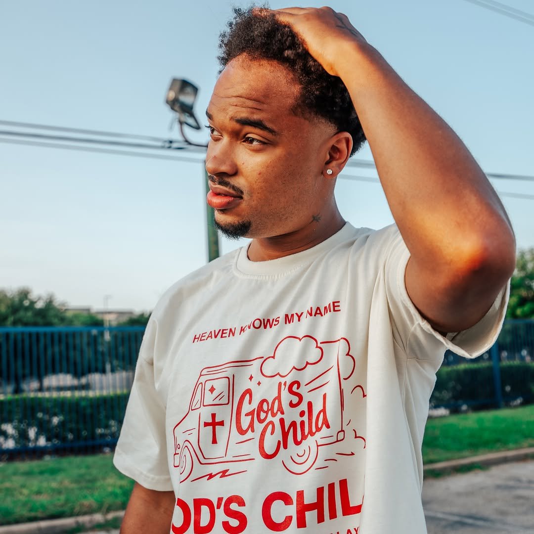 Jx.Zero with short curly hair, wearing a white T-shirt with red text and graphics, standing outdoors in front of a blue fence and power lines, touching his head with his right hand and looking to the side.