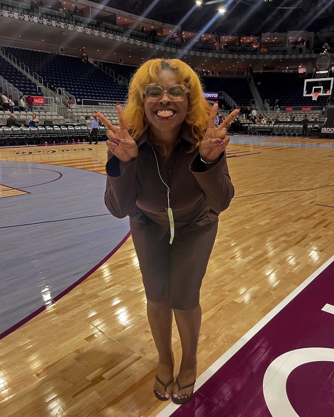 DĀHNI with curly blond hair and glasses on the Toronto Tempo basketball court making peace signs with both hands.