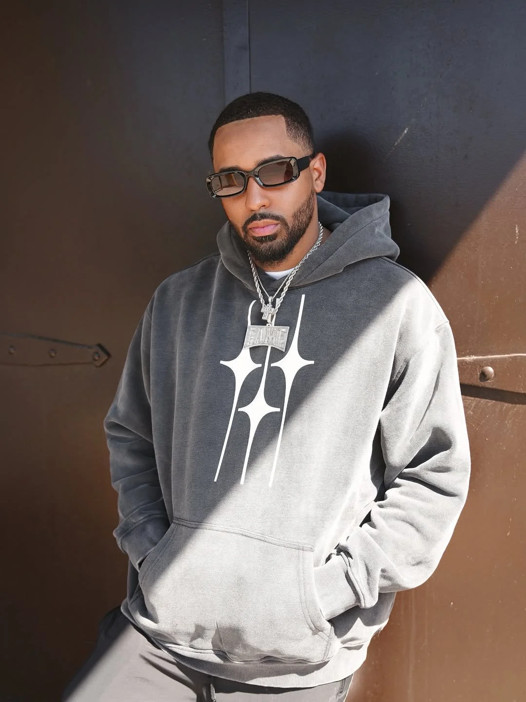 Fame Holiday wearing sunglasses, a gray hoodie with white graphic design, and layered necklaces, leaning against a dark wall.
