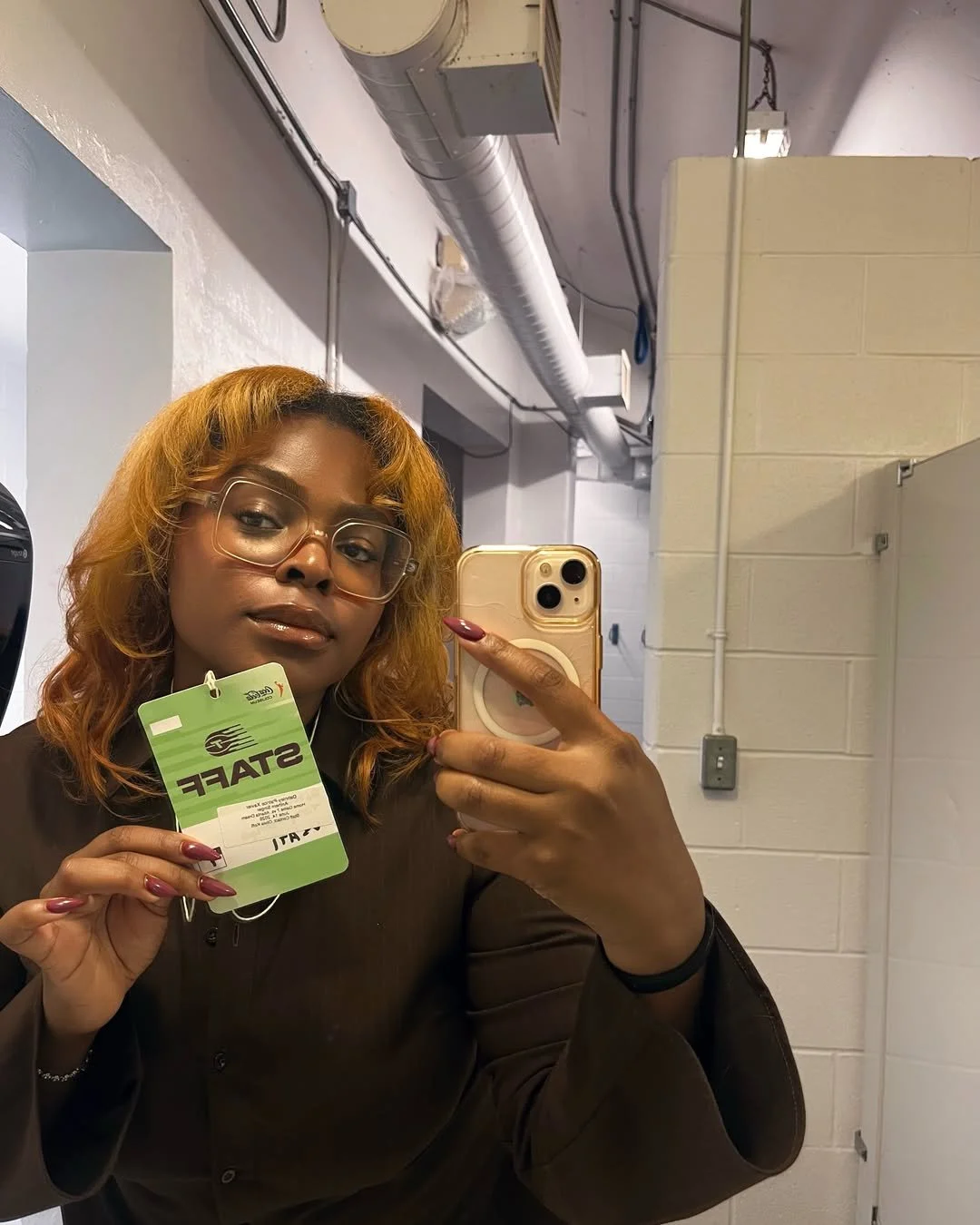  DĀHNI with orange hair and glasses taking a selfie in a restroom mirror, holding a green staff badge, wearing a dark brown shirt.
