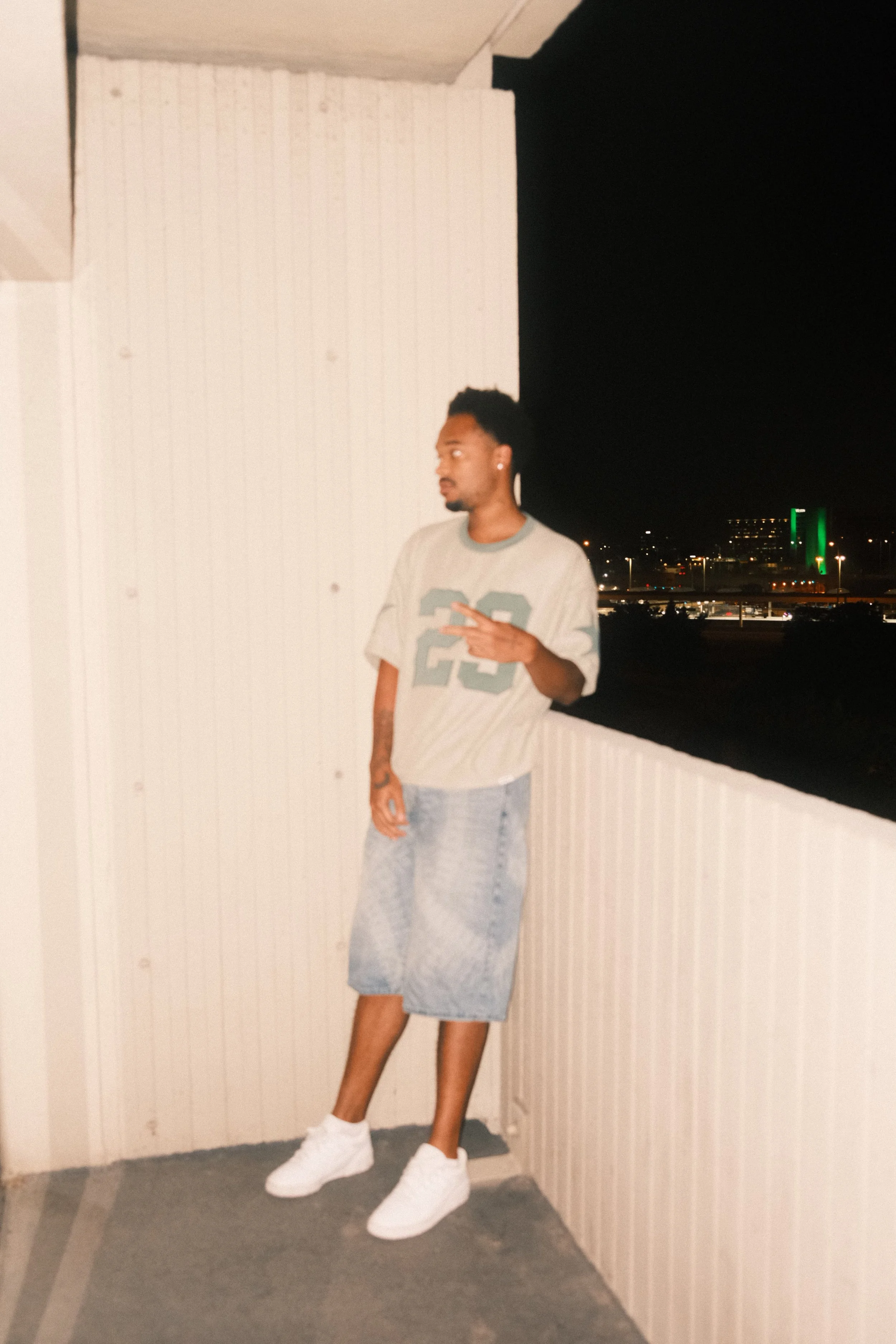 Jx.Zero with a beard and earrings posing at night on a balcony, wearing a gray T-shirt with large number 23 print, gray shorts, and white sneakers, with city lights and a green-lit building in the background.