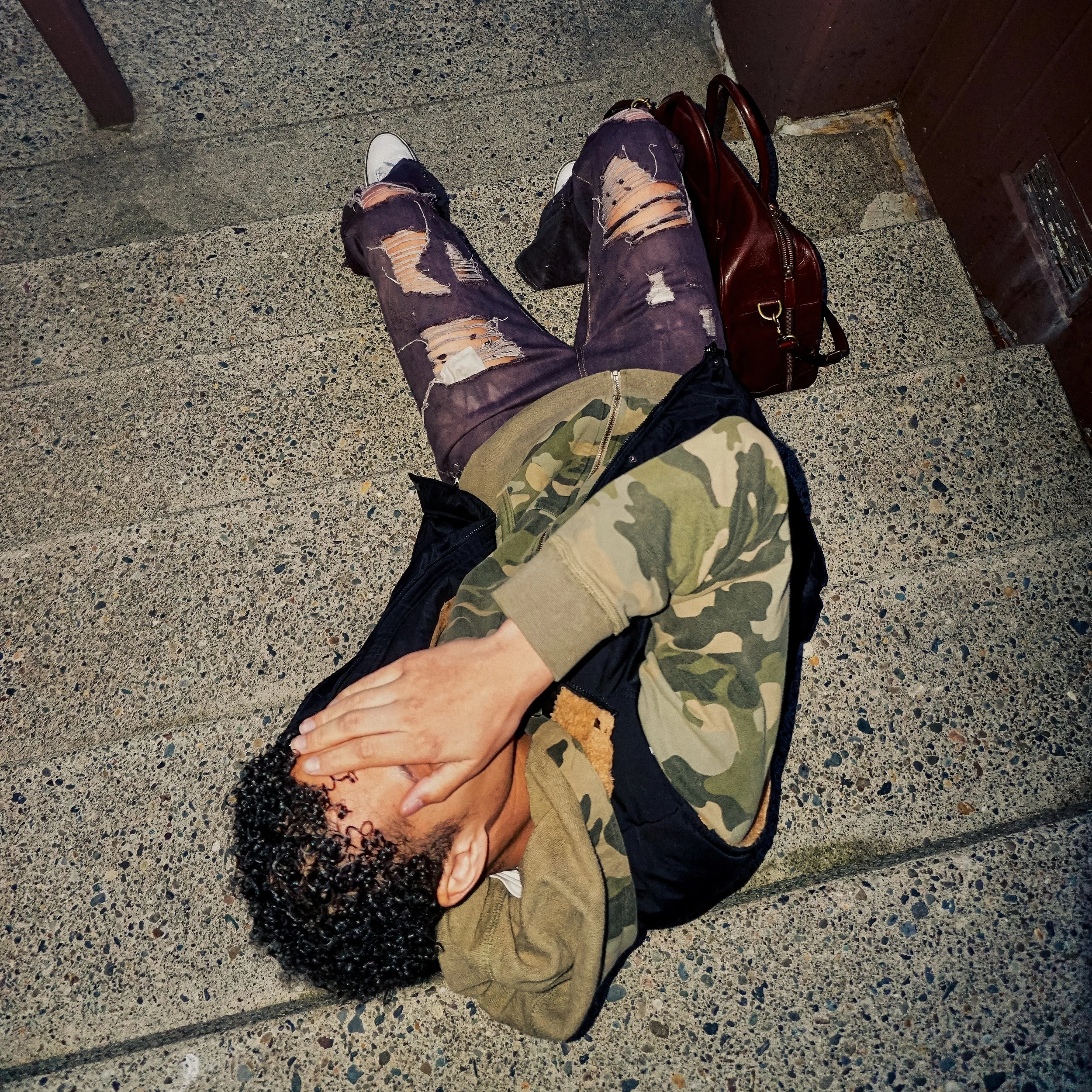 Jalen Jordan wearing ripped jeans, a camouflage jacket, and white sneakers sitting on concrete stairs with a brown leather bag nearby, holding hands with someone else.