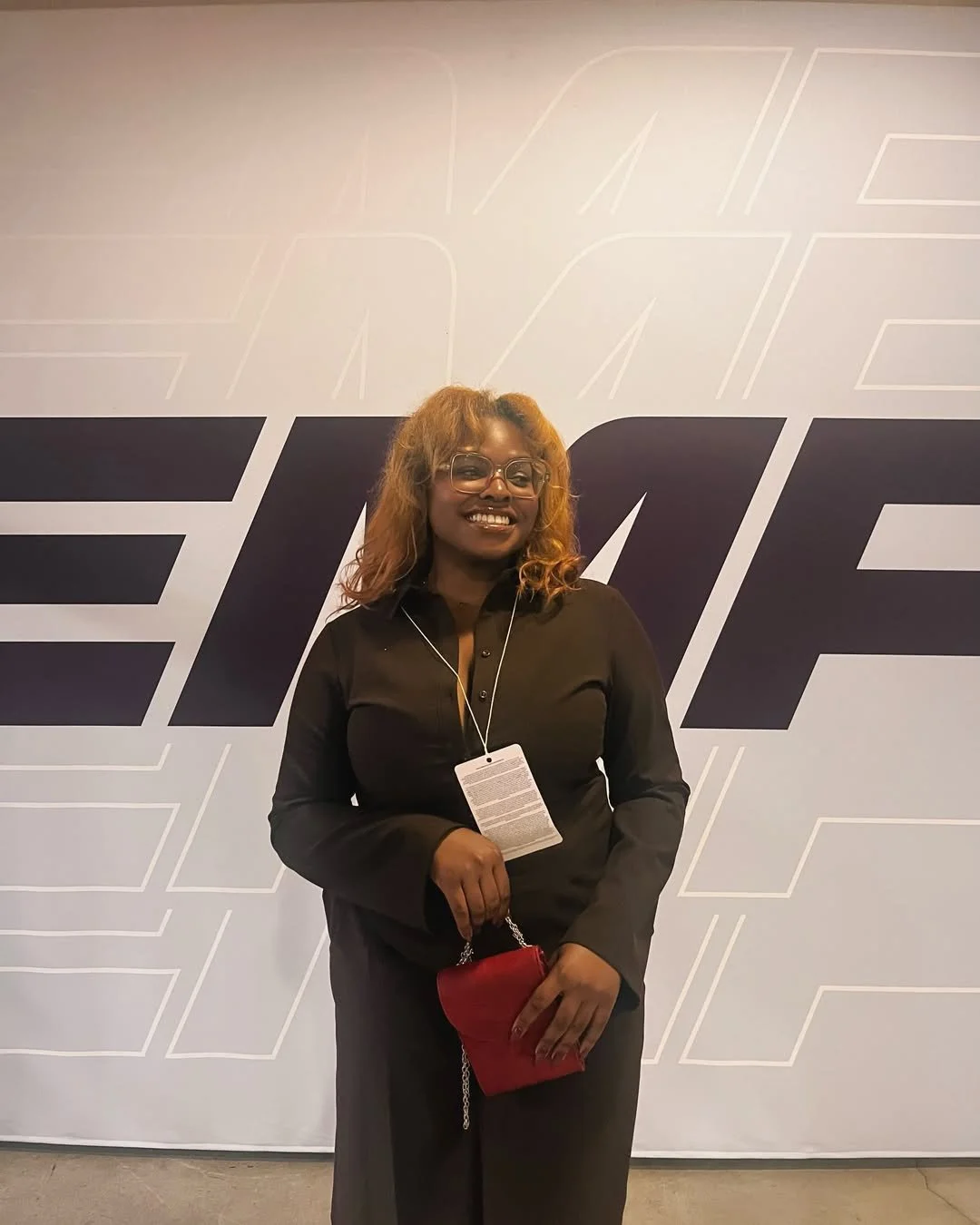  DĀHNI with glasses, curly hair, and a black outfit, smiling and holding a red purse, standing in front of a large wall with the word 'F1' in the background.