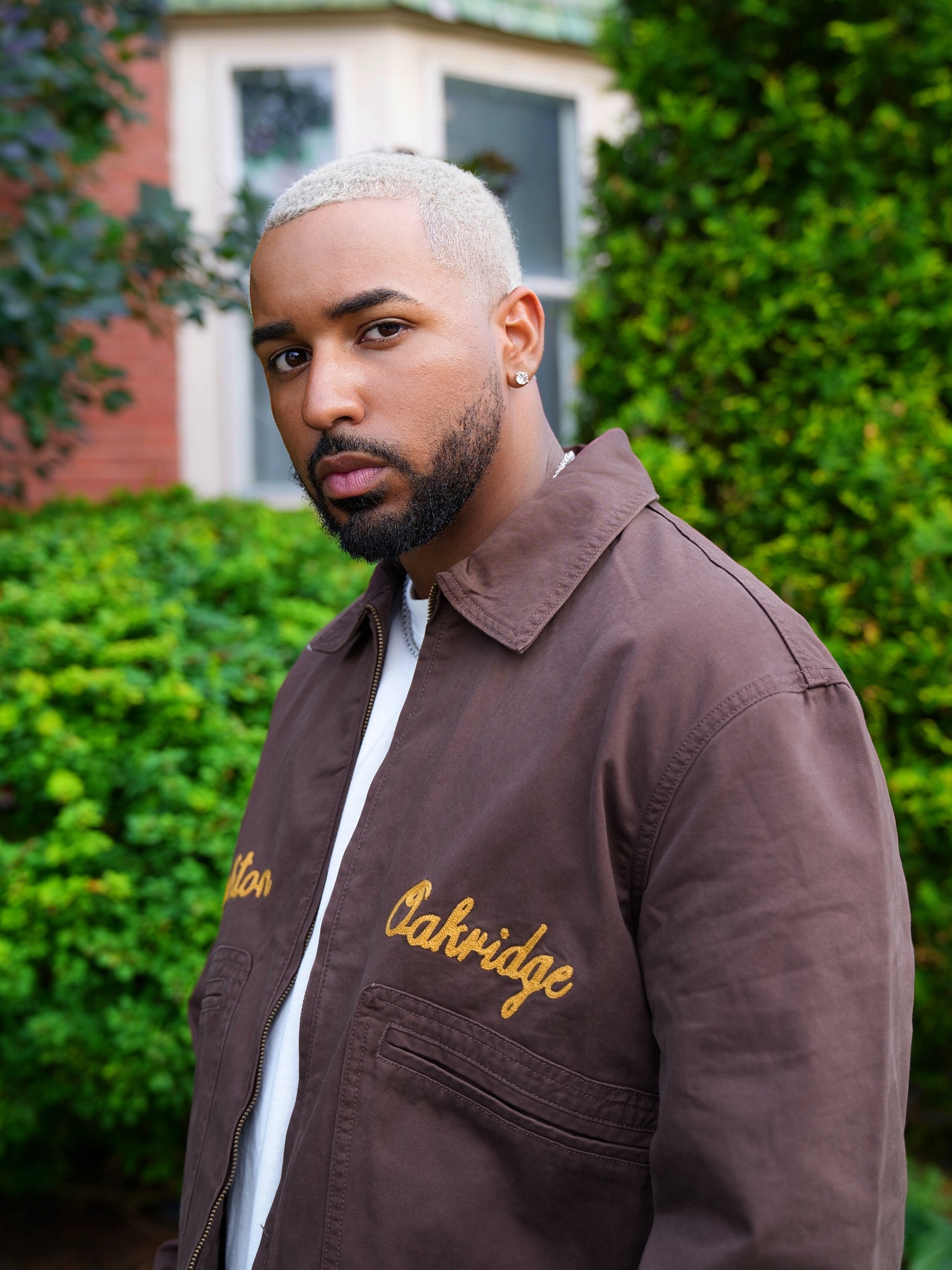 Fame Holiday with dyed blond hair and a beard stands outside with greenery and a house in the background, wearing a brown jacket with gold embroidery.
