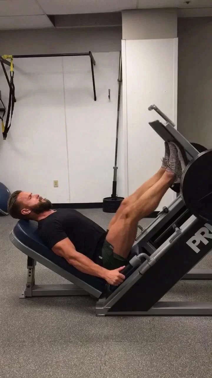 Man in gym performing squat exercise in front of dumbbell rack, wearing black shirt and yellow shoes.