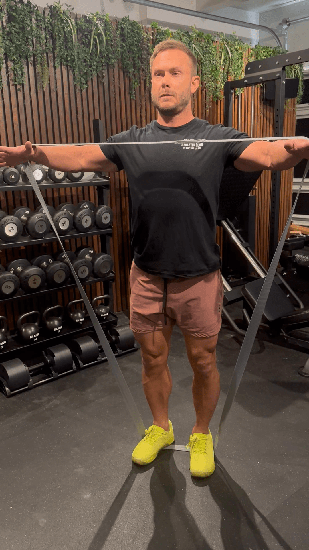 Man exercising with resistance band in gym, surrounded by weight racks, wearing a black shirt and pink shorts with neon yellow shoes.