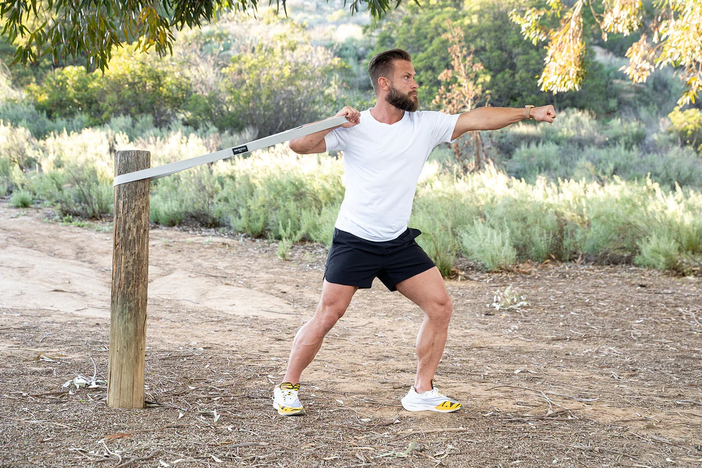 A man wearing a white t-shirt, black shorts, and white sneakers stretching his arms while using a resistance band attached to a post in an outdoor park setting.