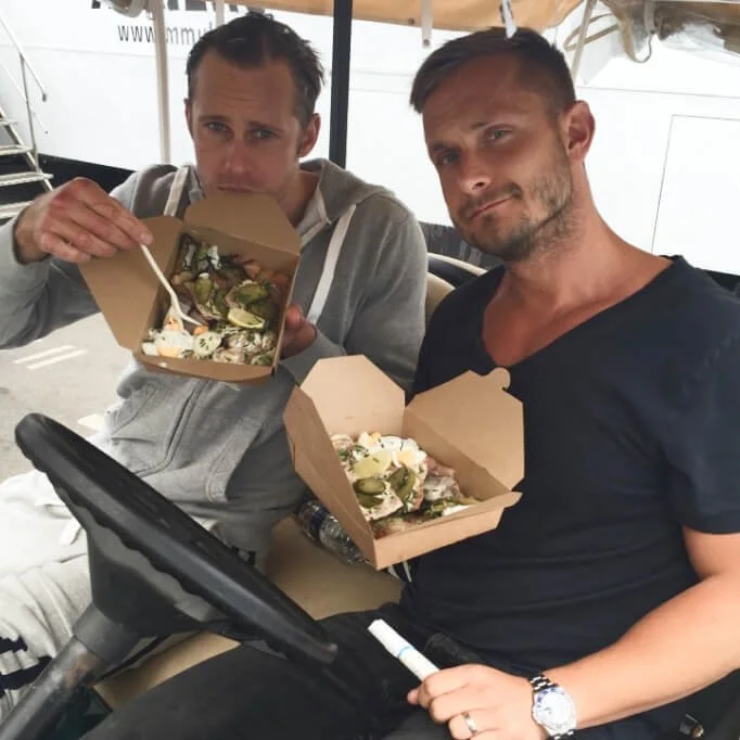 Two men seated together, each holding a takeout box filled with a mixed salad or food dish, sitting on a golf cart.