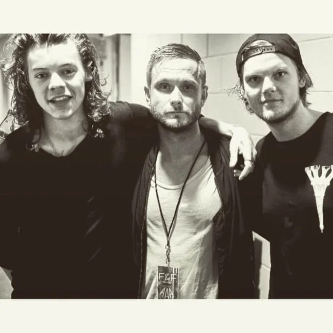 Three men posing together in a corridor, one wearing a concert pass lanyard.