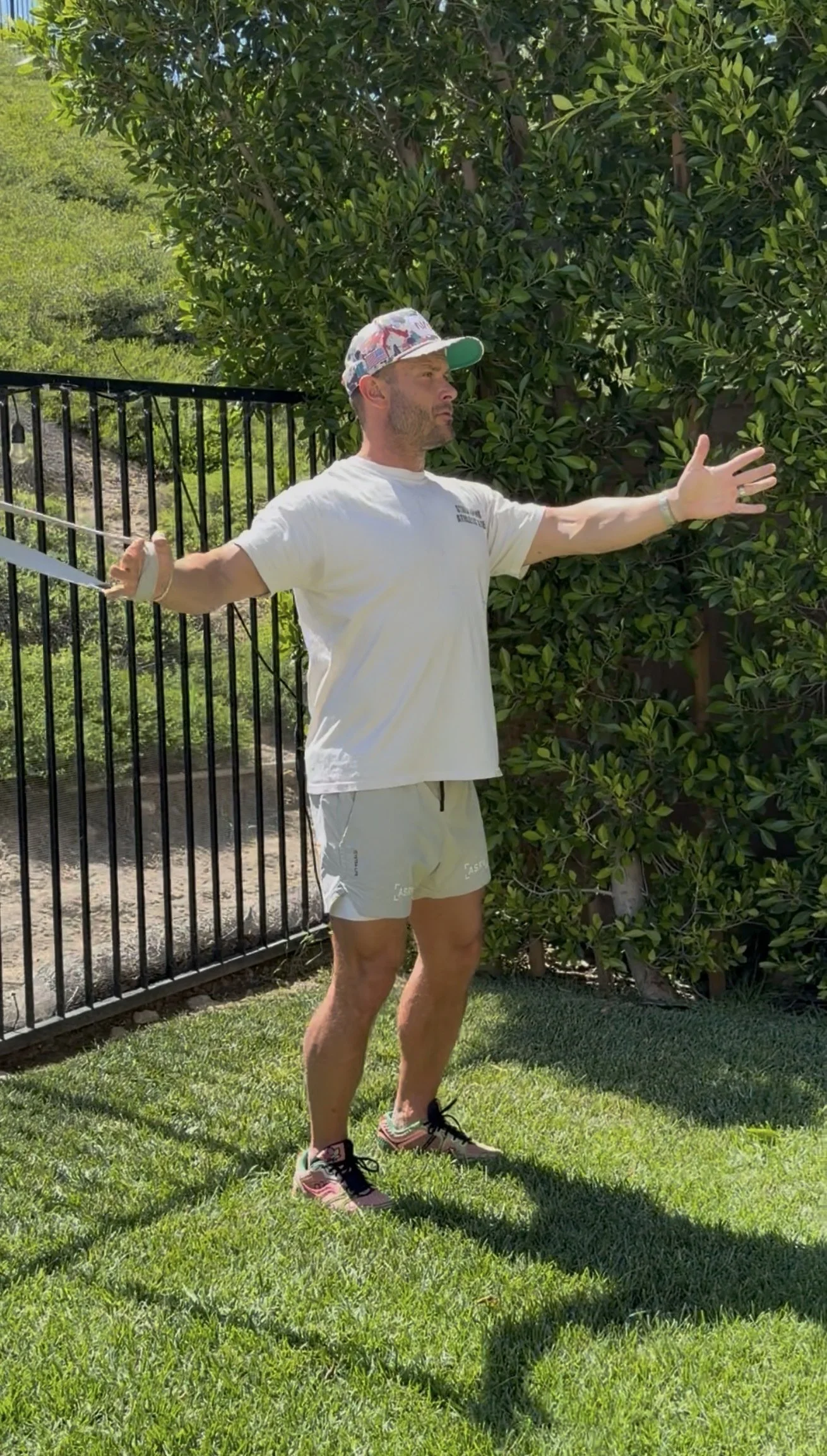 A man stretching outdoors on a grassy lawn with a fence and green bushes in the background, wearing a white T-shirt, light shorts, a floral baseball cap, and running shoes.