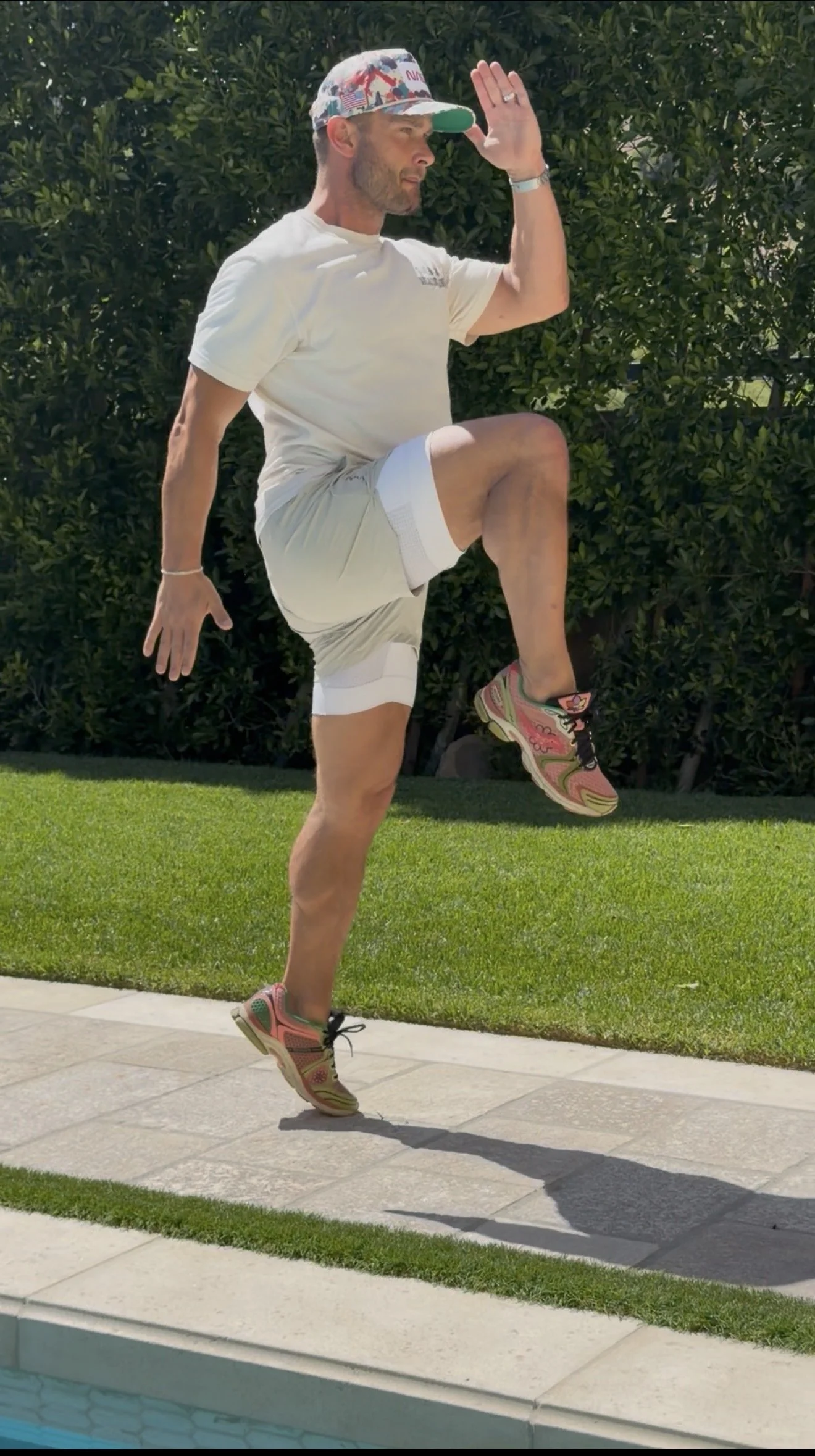 A man in a white t-shirt, shorts, and colorful sneakers is balancing on one leg while performing a yoga pose outside. He is holding his other leg up and is raising one hand to his forehead as if saluting. He is wearing a patterned cap and has a beard. There are green bushes and grass in the background.