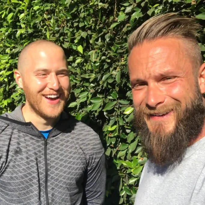 Two men smiling in front of a leafy green background outdoors.