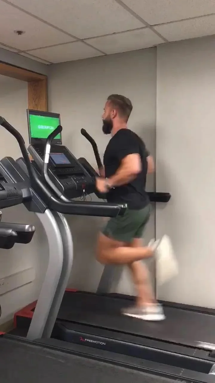 Man doing a squat exercise in a gym with dumbbells on a rack and wooden wall with plants.