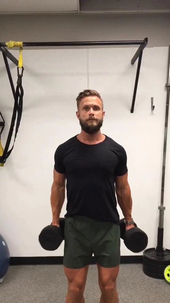 Man performing an exercise in a gym with dumbbells in the background, wearing a black shirt and green shoes.