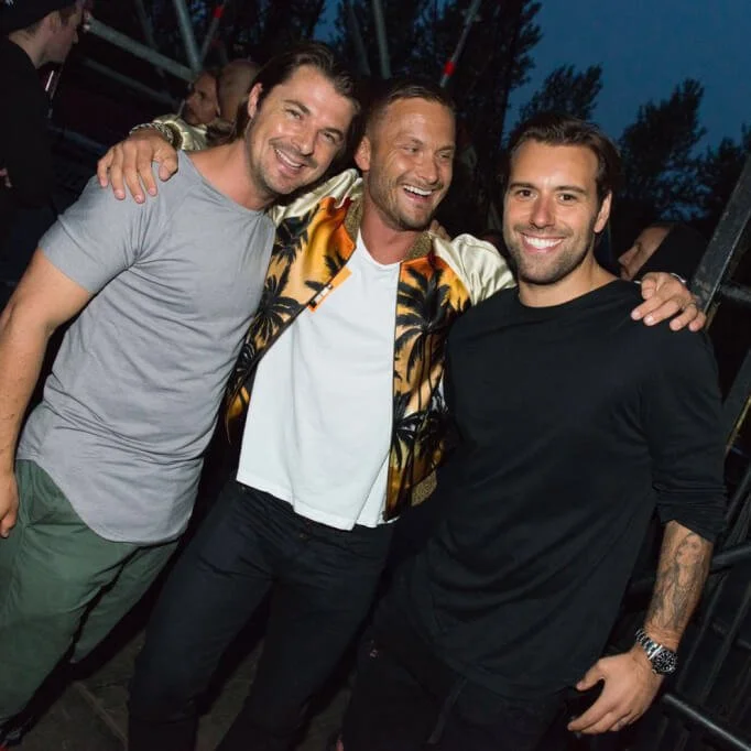 Three smiling men posing together at an outdoor event in the evening.