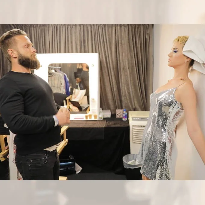 Man with beard posing playfully with a wax figure in a backstage dressing room, featuring a mirror and dressing table setup.