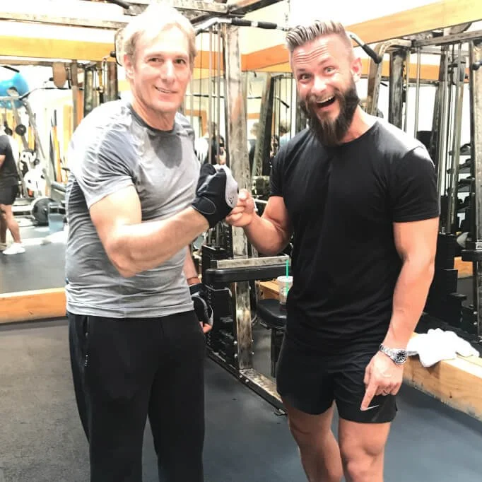 Two men shaking hands in a gym with exercise equipment in the background. One man is wearing a gray shirt and the other a black shirt, both are smiling.