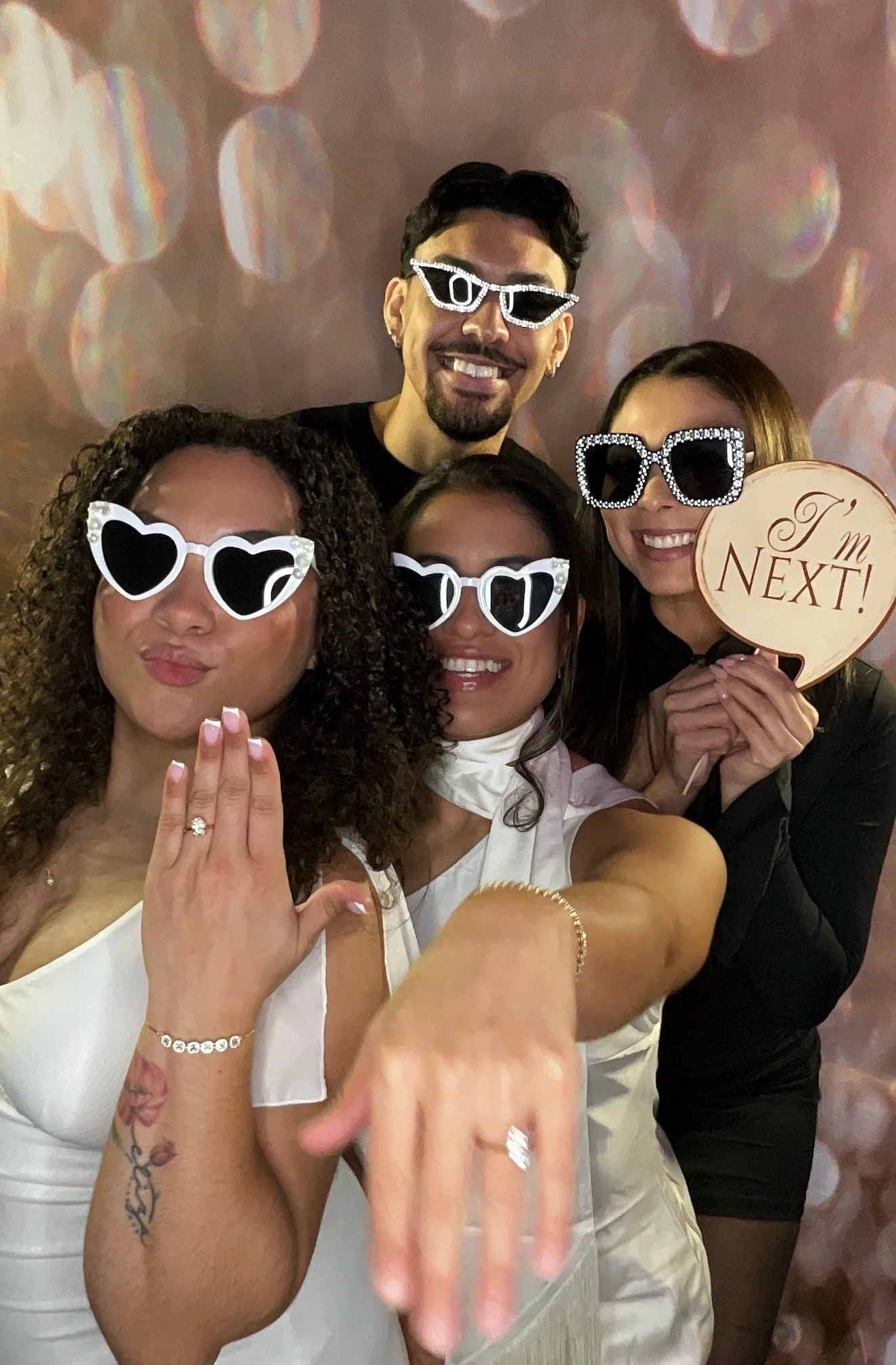 Group of five friends at a celebration, all wearing sunglasses and holding a speech bubble prop that says 'I'm next!' in front of a background with bokeh lights.