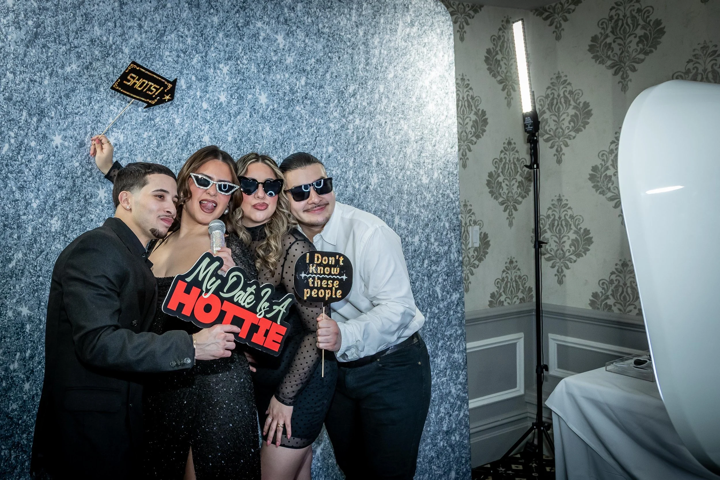 Four friends at a photo booth, wearing dark sunglasses, holding playful signs, and posing with a glittery backdrop with silver and blue tones. One holds a sign that says 'My Date is a HOTTE' and another a sign that says 'I Don't Know these people.' T