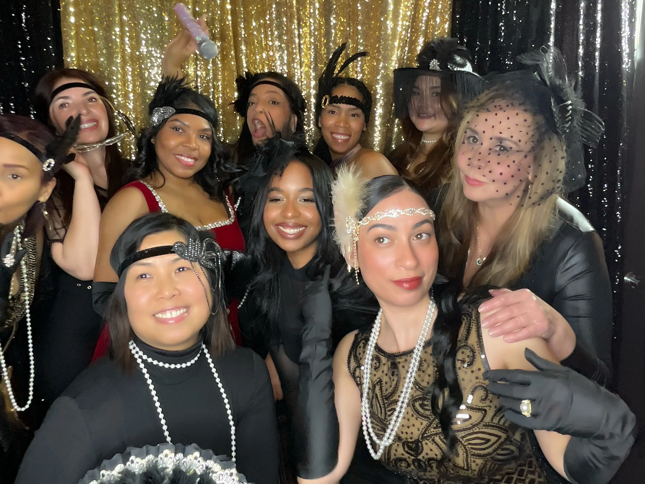 A group of women dressed in 1920s flapper costumes, wearing headbands with feathers, pearls, and vintage accessories, posing together in front of a gold and black sequin backdrop.