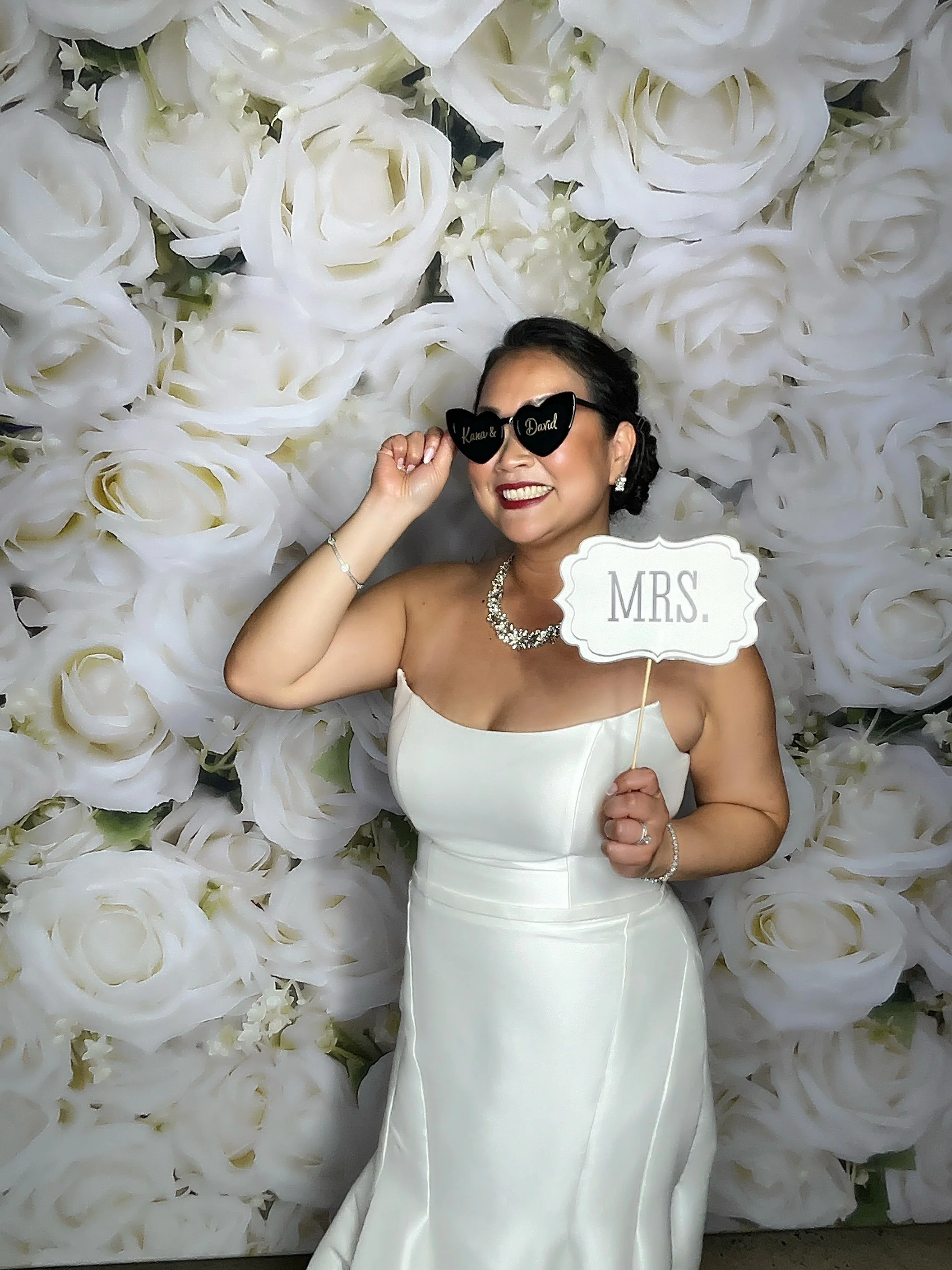 A woman in a white wedding dress is smiling, holding a sign that says 'MRS.', and adjusting heart-shaped sunglasses with the words 'Kama & David' on them. She is wearing jewelry and standing in front of a backdrop made of large white roses.