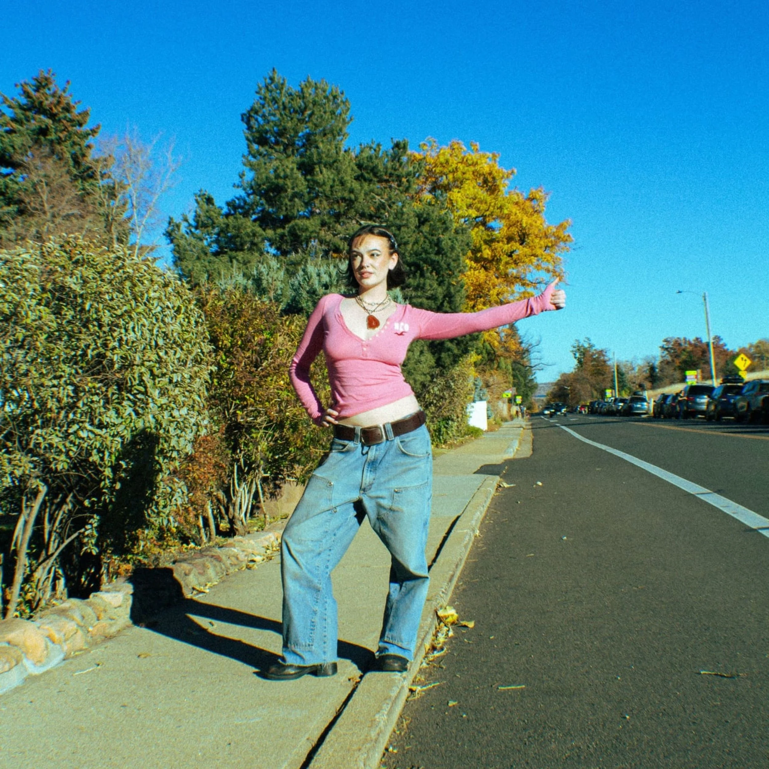 A woman standing on a sidewalk, wearing a pink long-sleeve shirt, baggy blue jeans, and a necklace, with her right hand on her hip and her left arm extended outward. She is outdoors near trees and parked cars on a clear, sunny day.