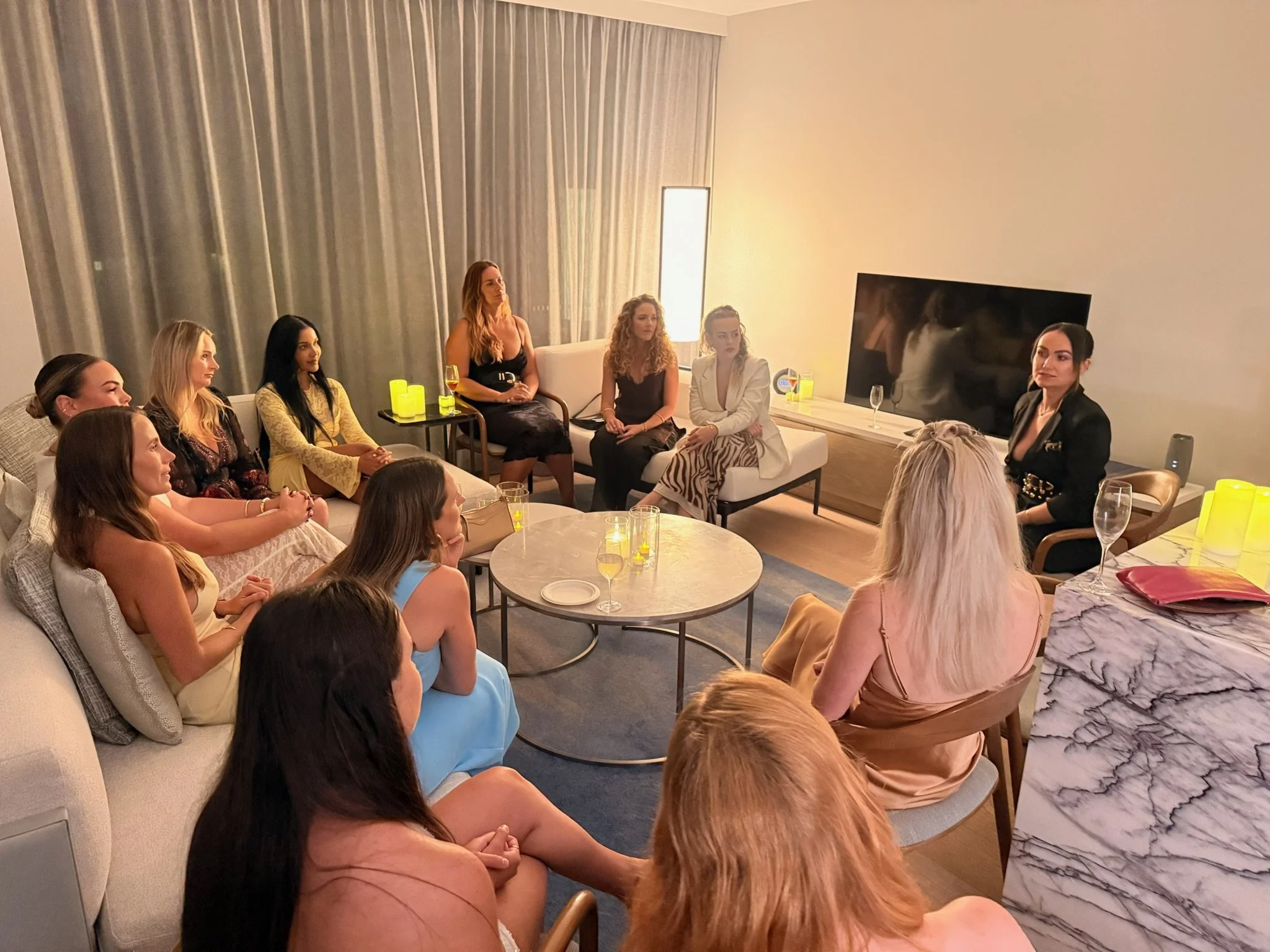 A group of women sitting on sofas and chairs in a living room, watching a woman standing in front of them. The room has beige walls, gray curtains, a large TV on the wall, and a marble table with drinks and candles.
