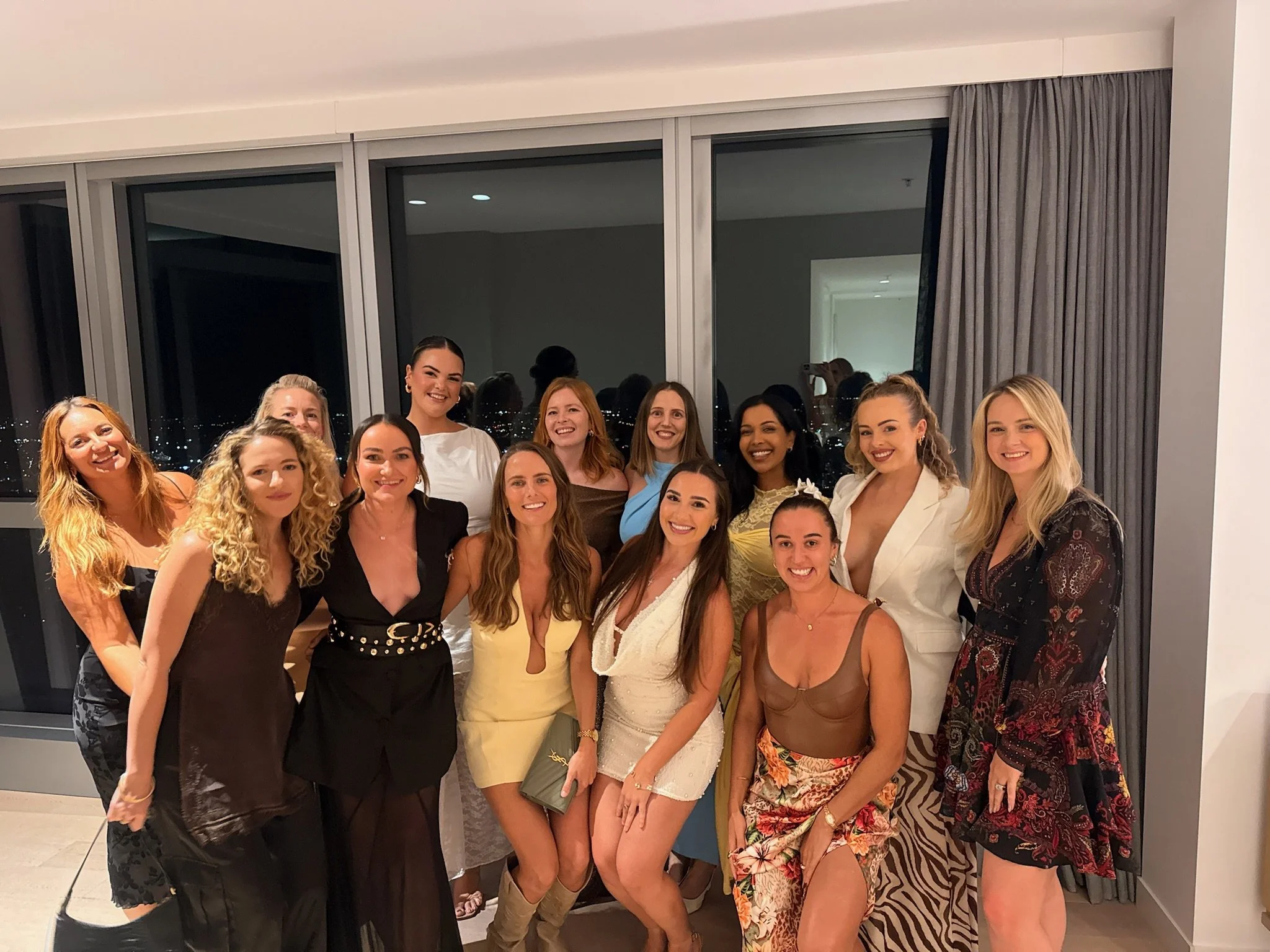 A group of women dressed in formal and stylish clothes posing together indoors, with a large window and city lights visible in the background.