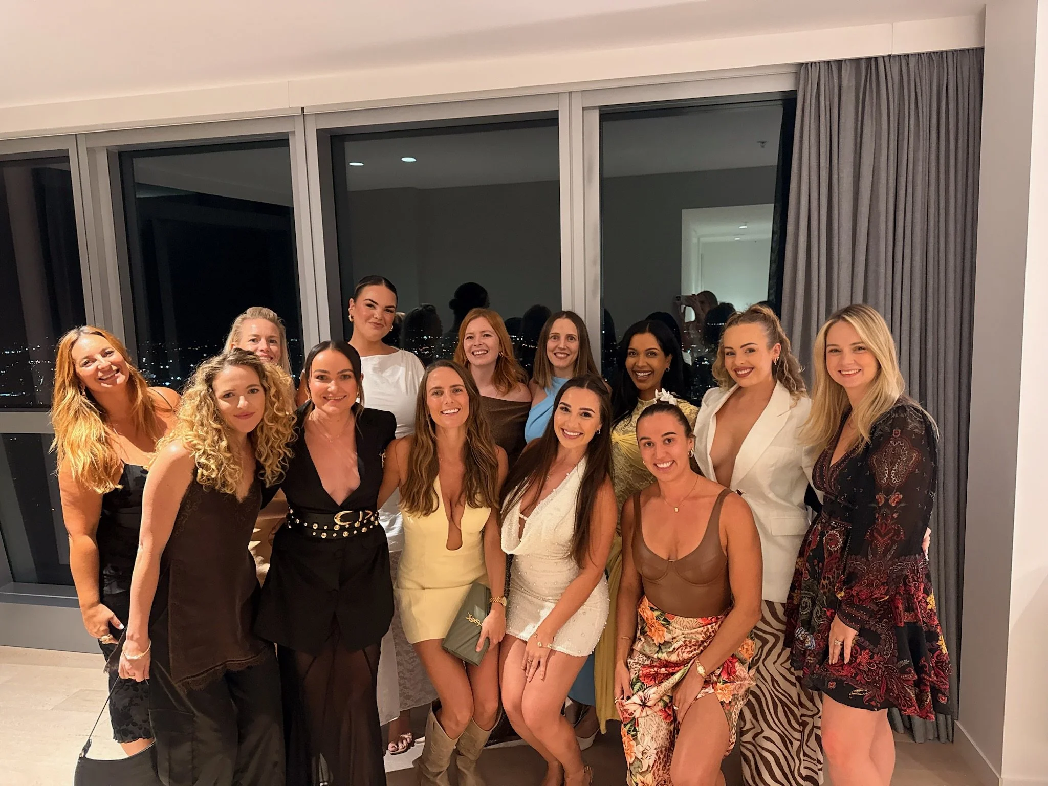 A group of women dressed in formal attire gathered together indoors for a photo, with a city view visible through large windows in the background.