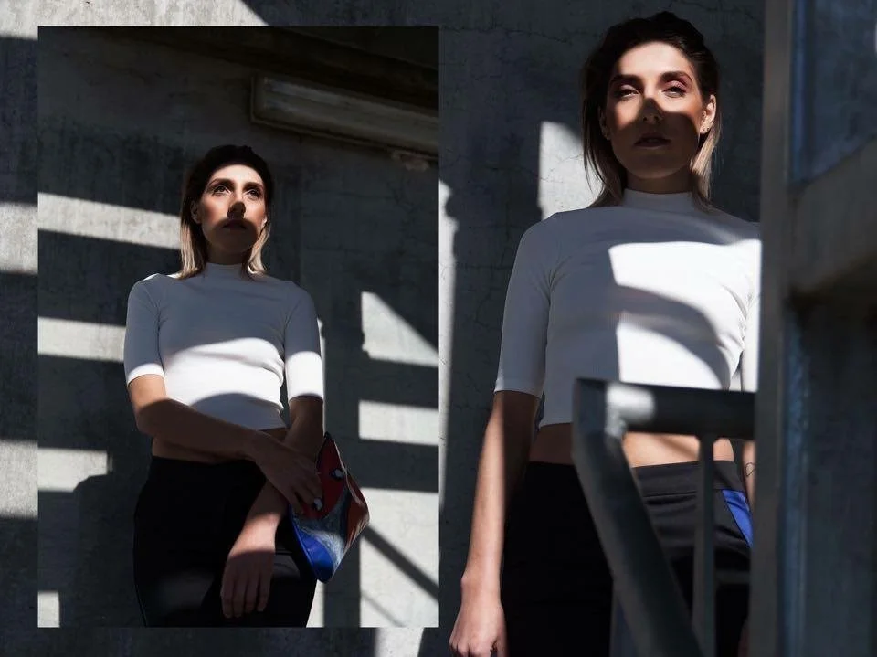 A woman in a white top standing against a concrete wall with striped shadows, holding a colorful clutch, with sunlight highlighting her face and shadow patterns.