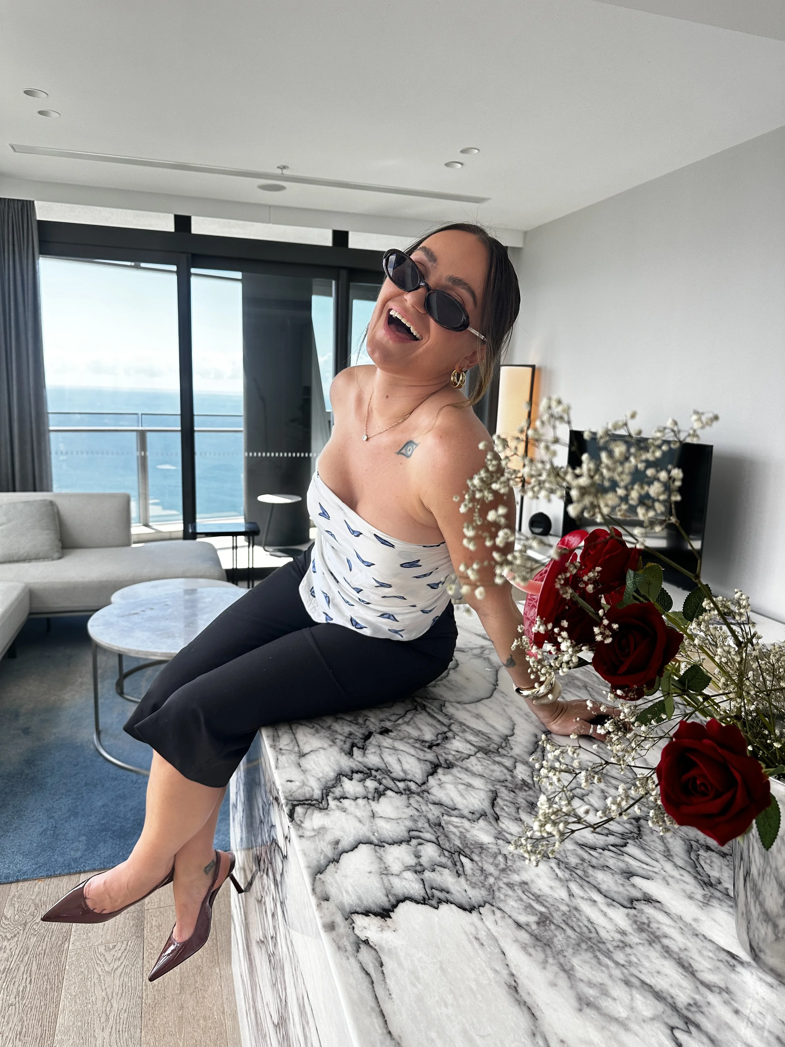 Woman in sunglasses and a sleeveless top smiling and leaning on a marble counter, with a bouquet of red and white flowers, in a modern living room with large windows and view of the ocean.
