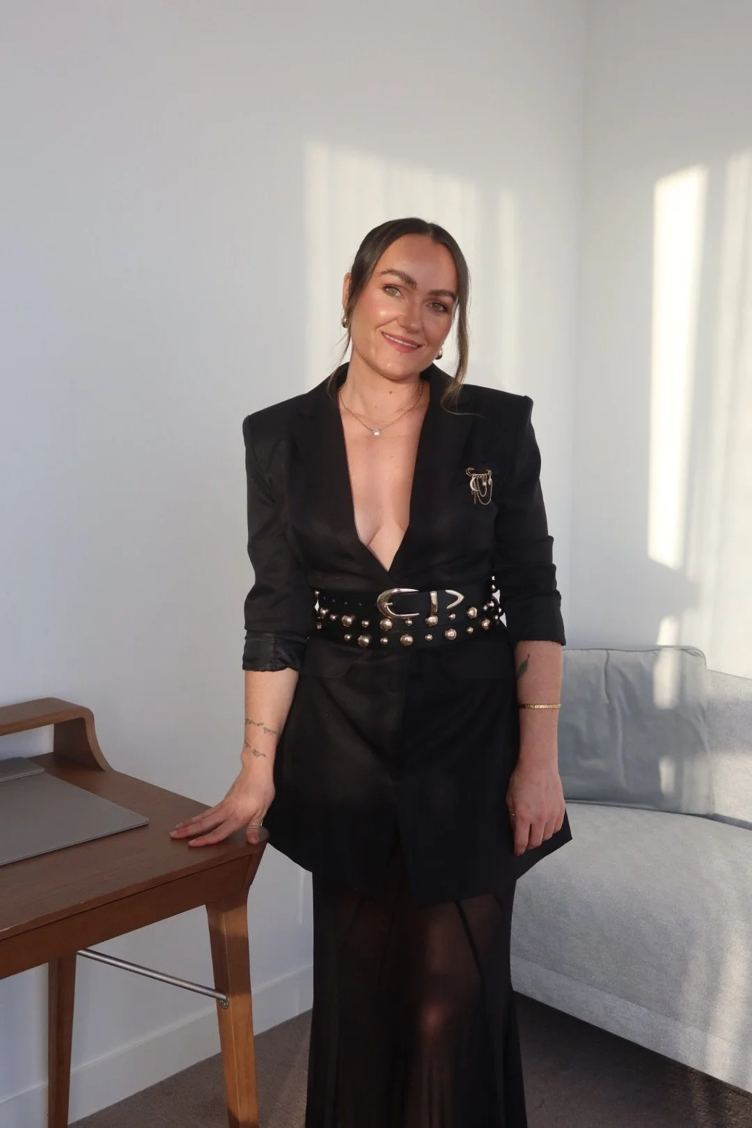 A woman with dark hair and light skin, dressed in a black blazer with a deep neckline, adorned with a brooch, wearing a studded black belt and black sheer skirt, standing in a room with white walls, sunlight coming through windows, a gray sofa, and a wooden desk with a laptop.