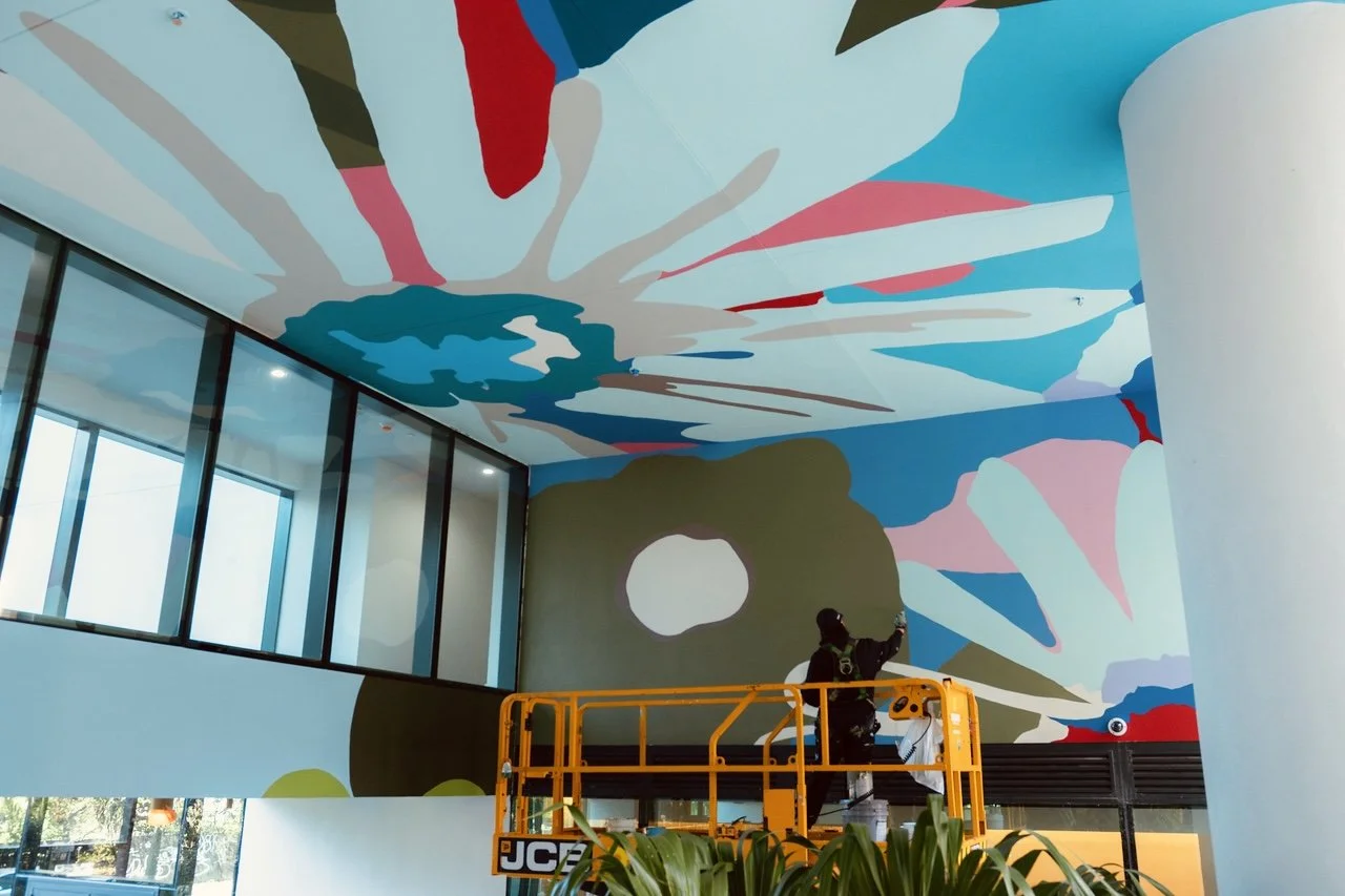 Worker on a yellow lift installing a colorful abstract mural on a large wall and ceiling.