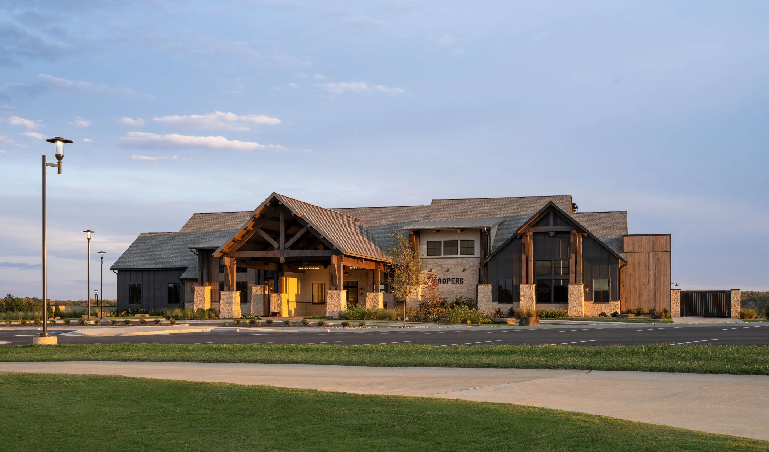 A modern building with a mix of stone, dark wood, and light siding, featuring a gabled roof and large windows, surrounded by a landscaped area with small trees and street lamps, under a partly cloudy sky.