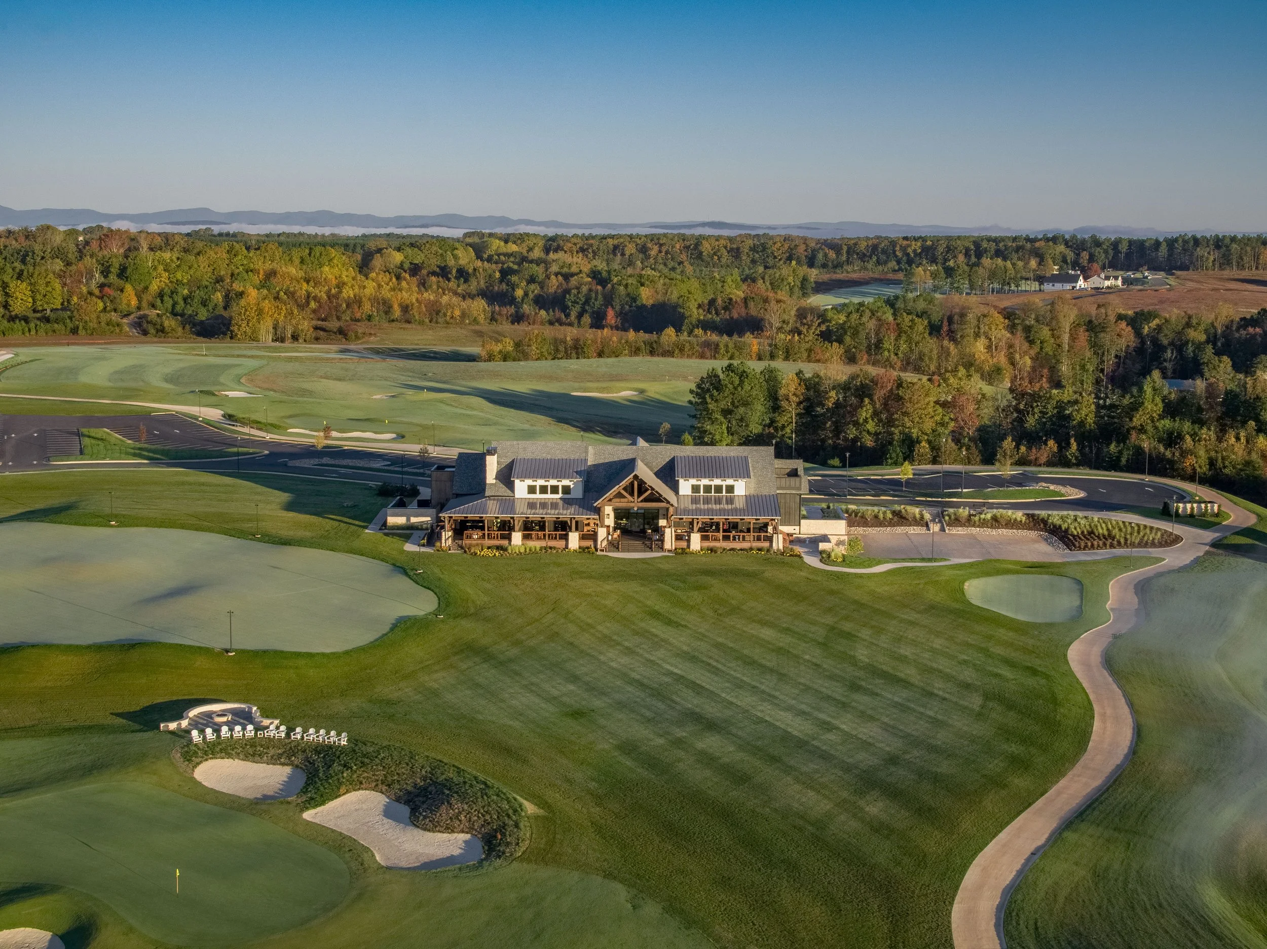 An aerial view of a golf course surrounded by trees and a few buildings. The course has sand bunkers and a parking lot nearby. In the background, there are mountains and a clear blue sky.