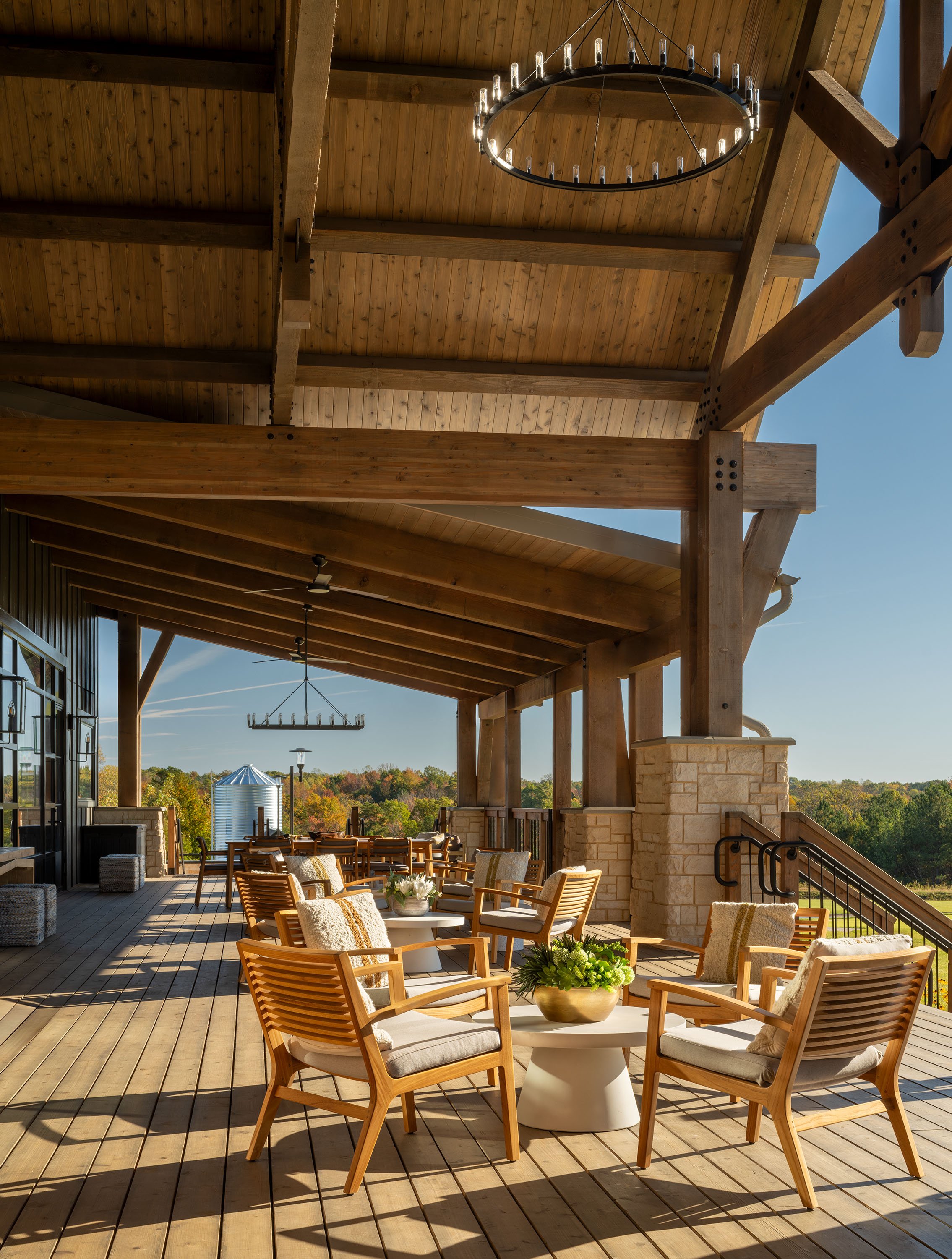Outdoor patio area with wooden furniture, chairs, and a table with a plant. Wooden ceiling with beams and a chandelier, with clear blue sky and trees in the background.
