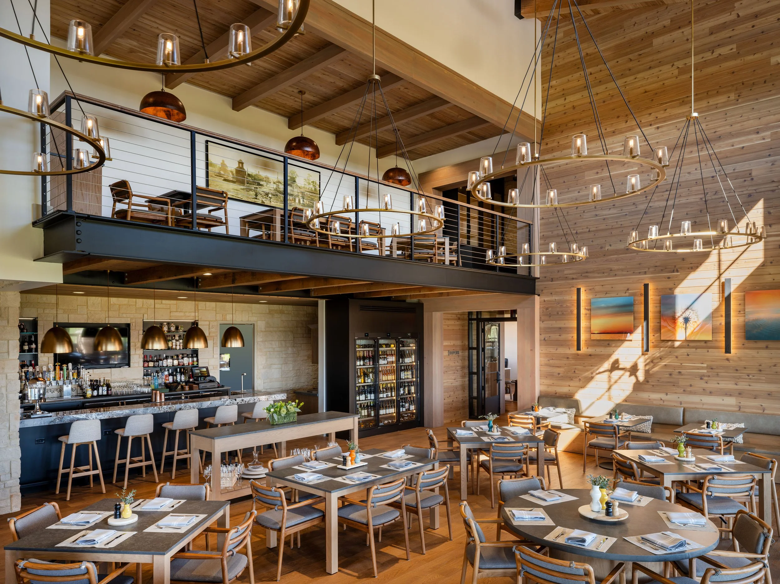 Interior of a restaurant with wooden ceiling and walls, modern chandeliers, a balcony with chairs and a large framed painting, a bar area with bottles and glasses, and tables set with napkins, cutlery, and small flower arrangements.