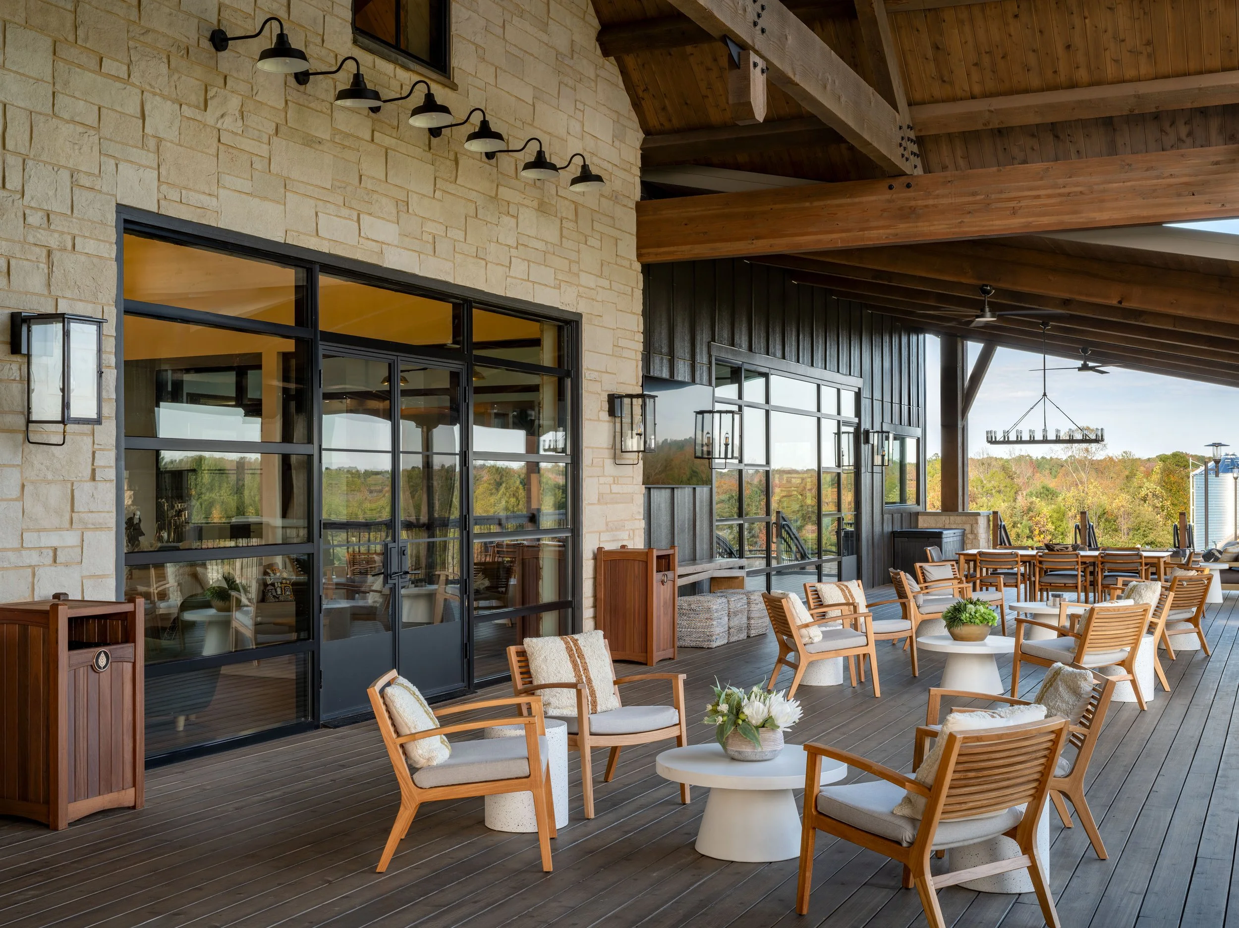 An outdoor patio with wooden chairs and tables decorated with flowers, large windows, wall-mounted lights, and a roof with wooden beams. In the background, there are trees and a blue sky.