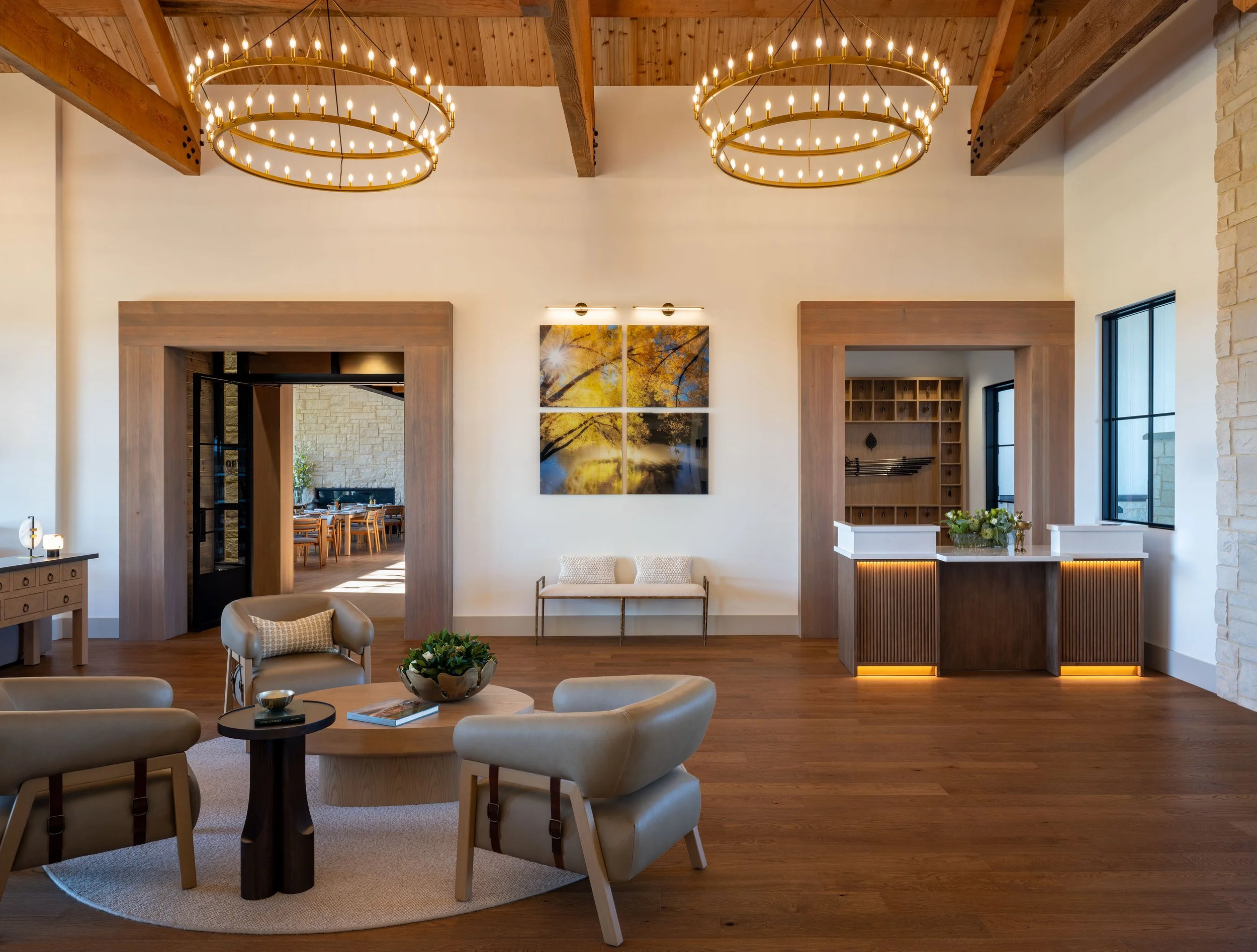 Living room with beige armchairs around a round black coffee table, wooden floors, modern decor, a large landscape painting, and brass chandelier lights.