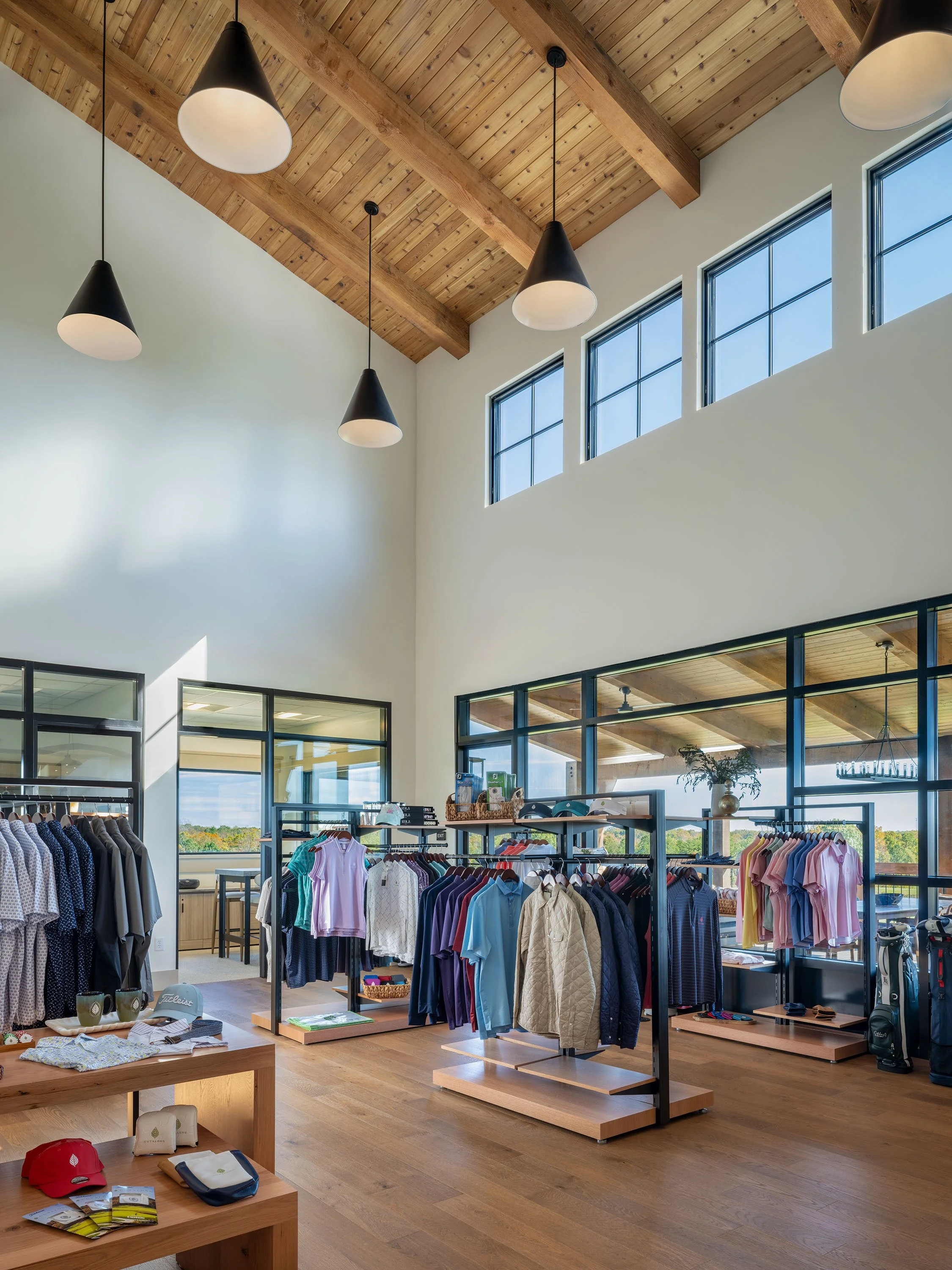 Interior of a clothing store with wooden flooring, high ceiling, large windows, and hanging lights, displaying various apparel on racks.