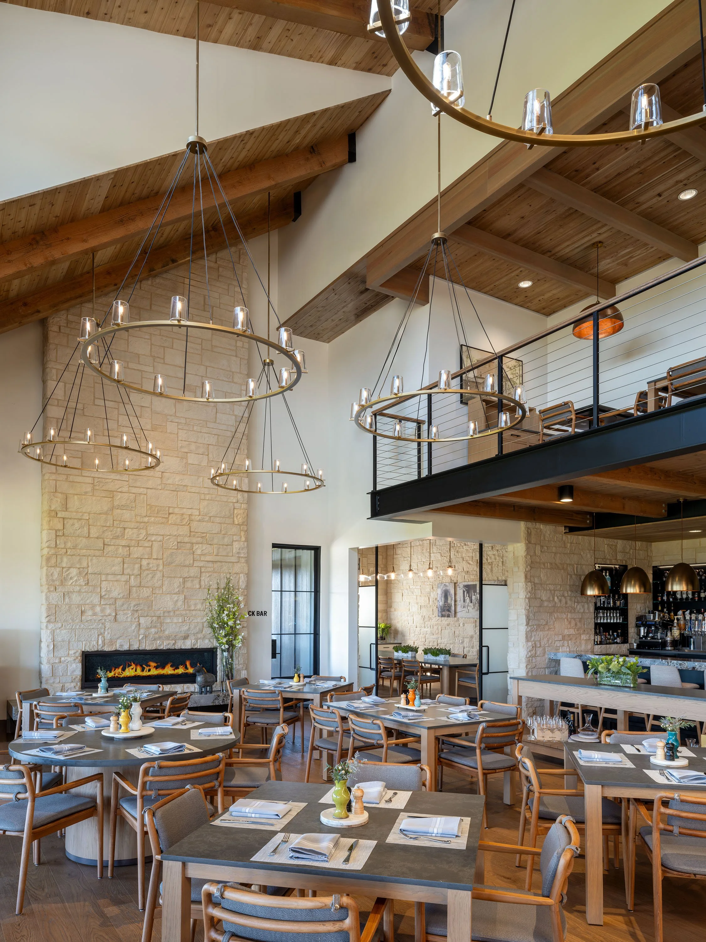 A spacious restaurant interior with wooden beams and a high ceiling, featuring multiple circular chandeliers, tables set with napkins, silverware, and small decorative items, a stone fireplace, and a balcony with a black railing.