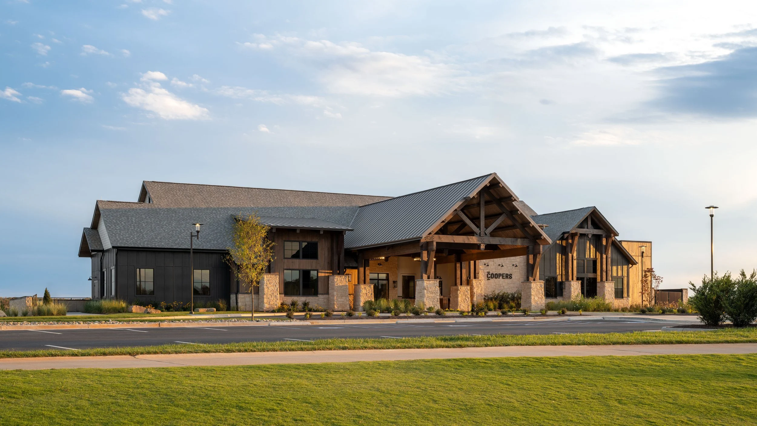 Exterior of a large building with wood and stone accents, a gabled roof, and sign that reads 'Coopers', situated along a street with parking spaces and lampposts under a cloudy sky.