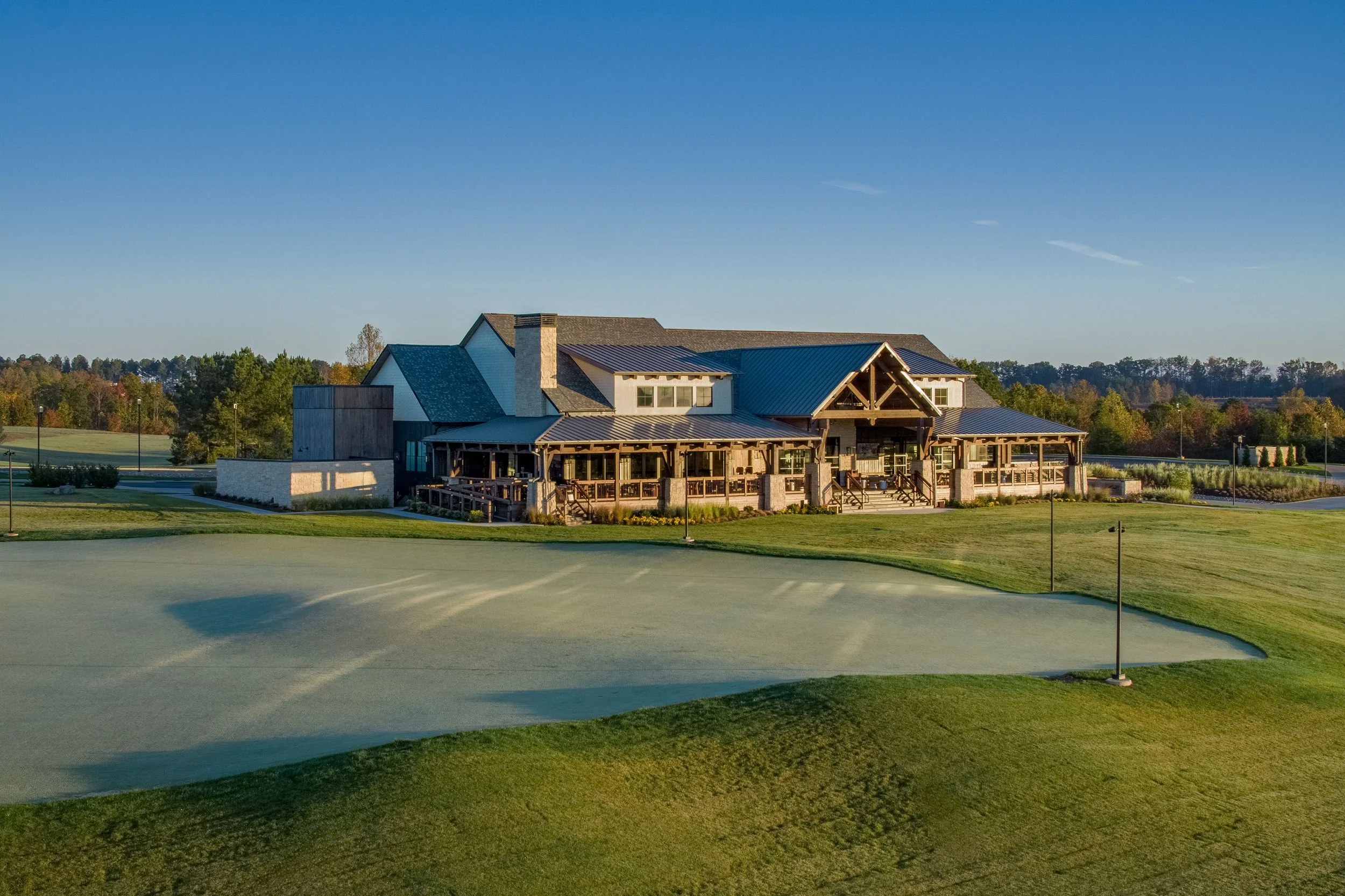 A large building with a rustic design, featuring a metal roof, wooden beams, and stone accents, situated beside a golf course or sports field with grass and a pathway, surrounded by trees and a clear blue sky.