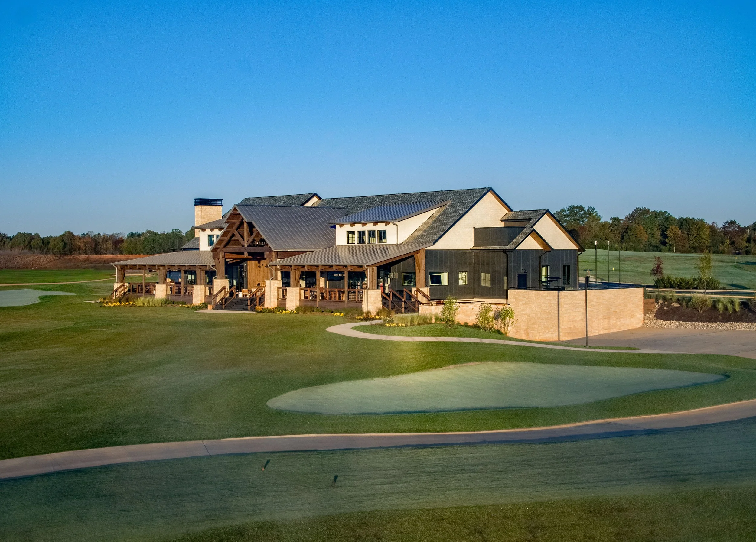 A large house with a modern design, featuring a combination of wood and black exterior materials, situated in a well-kept grassy area. The house has a prominent porch with wooden beams and steps leading up to the entrance, and a chimney on one side. 