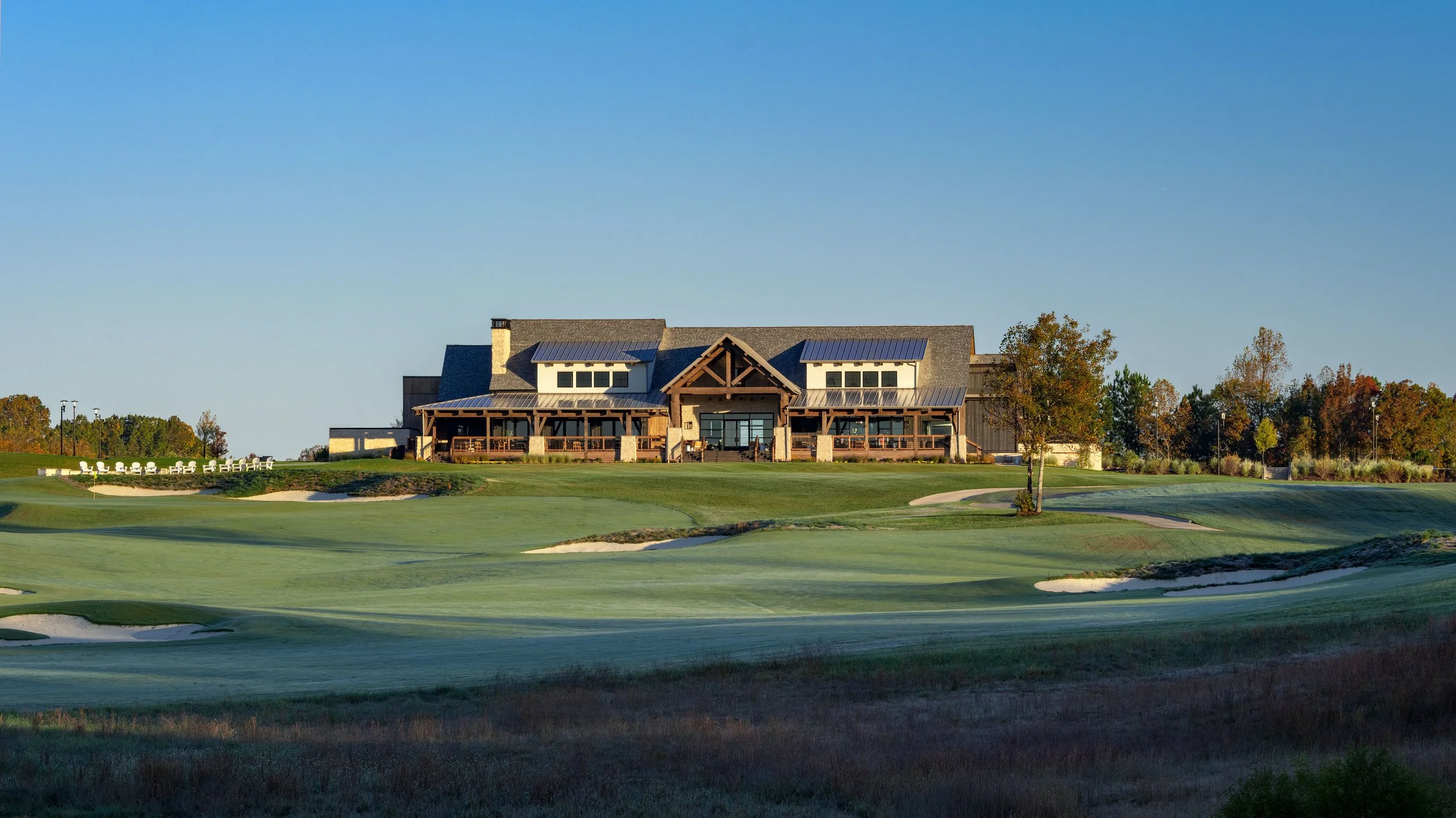 A large clubhouse at a golf course surrounded by green fairways, sand traps, and trees with autumn foliage under a clear blue sky.