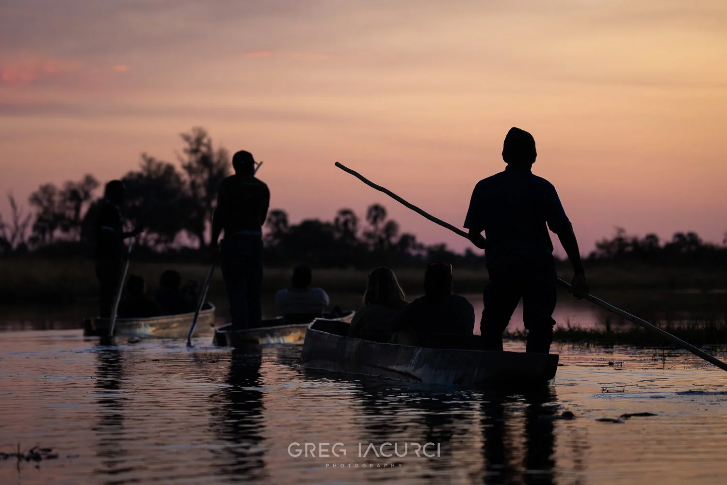 Men stand in canoe-like boats with poles during a pink sunset.